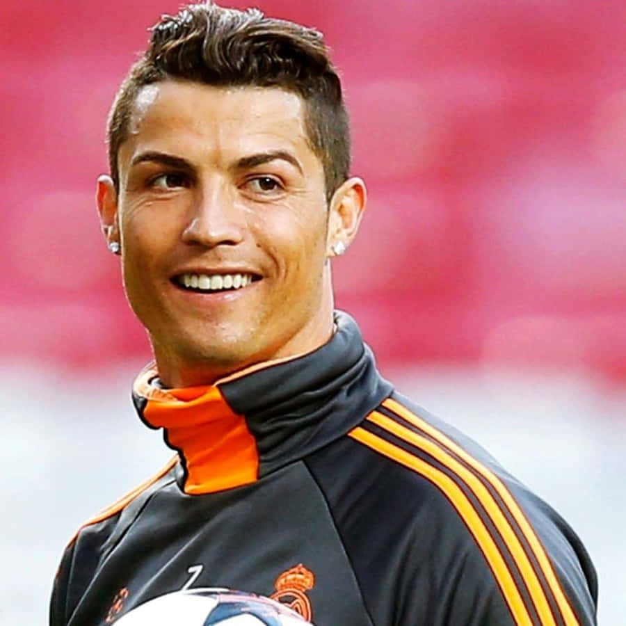 Ronaldo 桌布 壁紙 — 一位面帶微笑的足球員身穿黑色和橙色的訓練服，在模糊的紅色體育場背景下抱著一個足球。