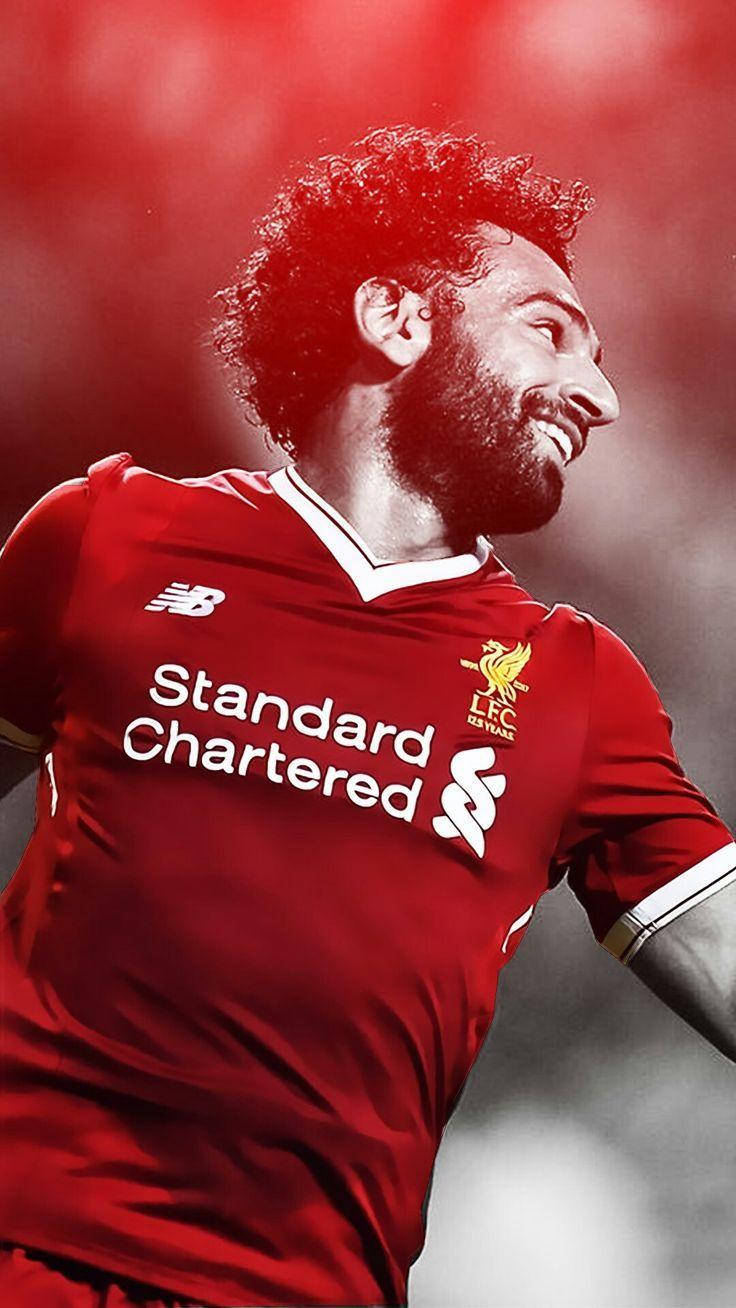 Salah 桌布 壁紙 — 一位面帶微笑的足球運動員身穿紅色利物浦球衣，在模糊的紅色背景下以強烈的喜悅慶祝。