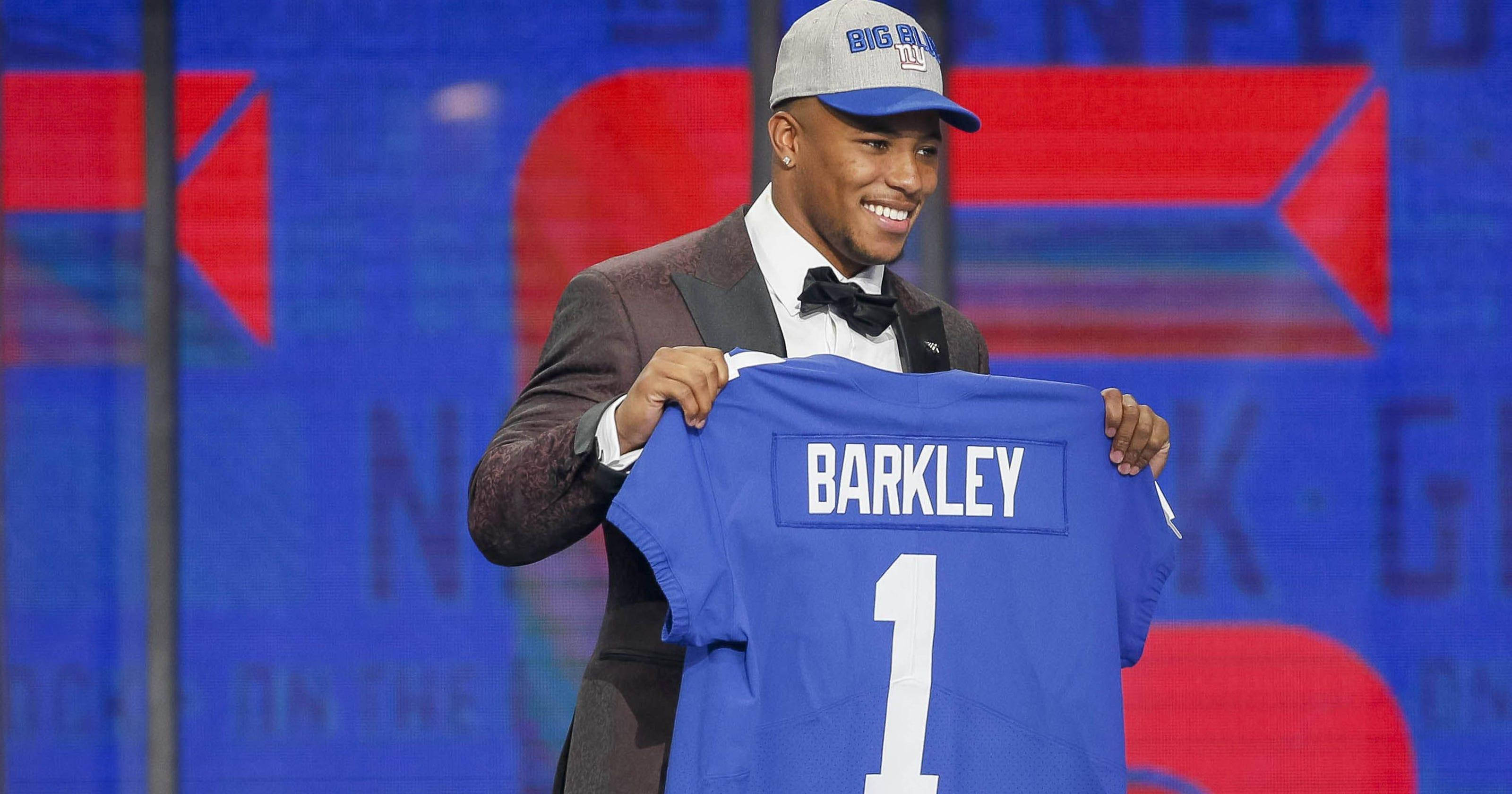 Saquon Barkley 桌布 壁紙 — 一位面帶微笑的運動員身穿西裝和帽子，在舞台上自豪地舉起一件印有「BARKLEY」和號碼「1」的藍色美式足球球衣。