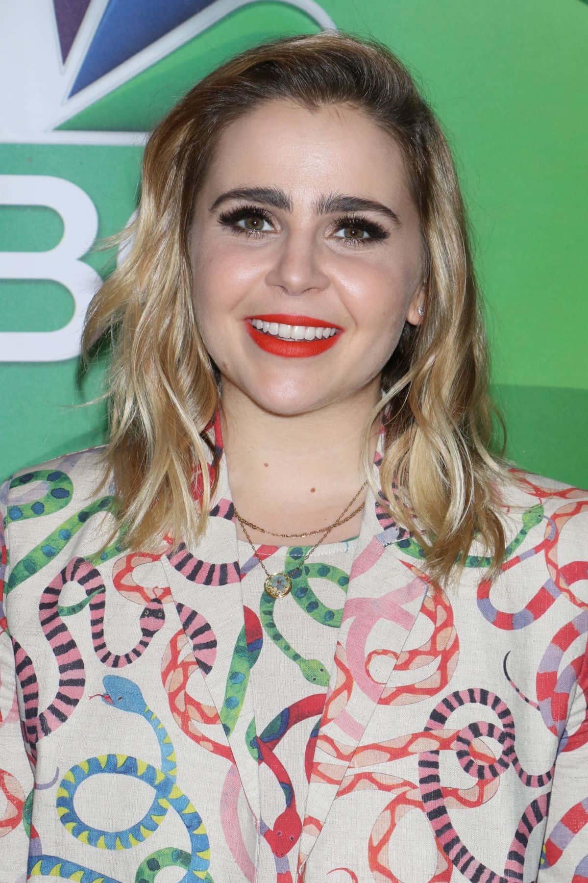 Mae Whitman 桌布 壁紙 — 一位面帶微笑的金髮女子，塗著大膽的紅色唇膏，穿著色彩繽紛的蛇紋西裝外套，背景是綠色的NBC。
