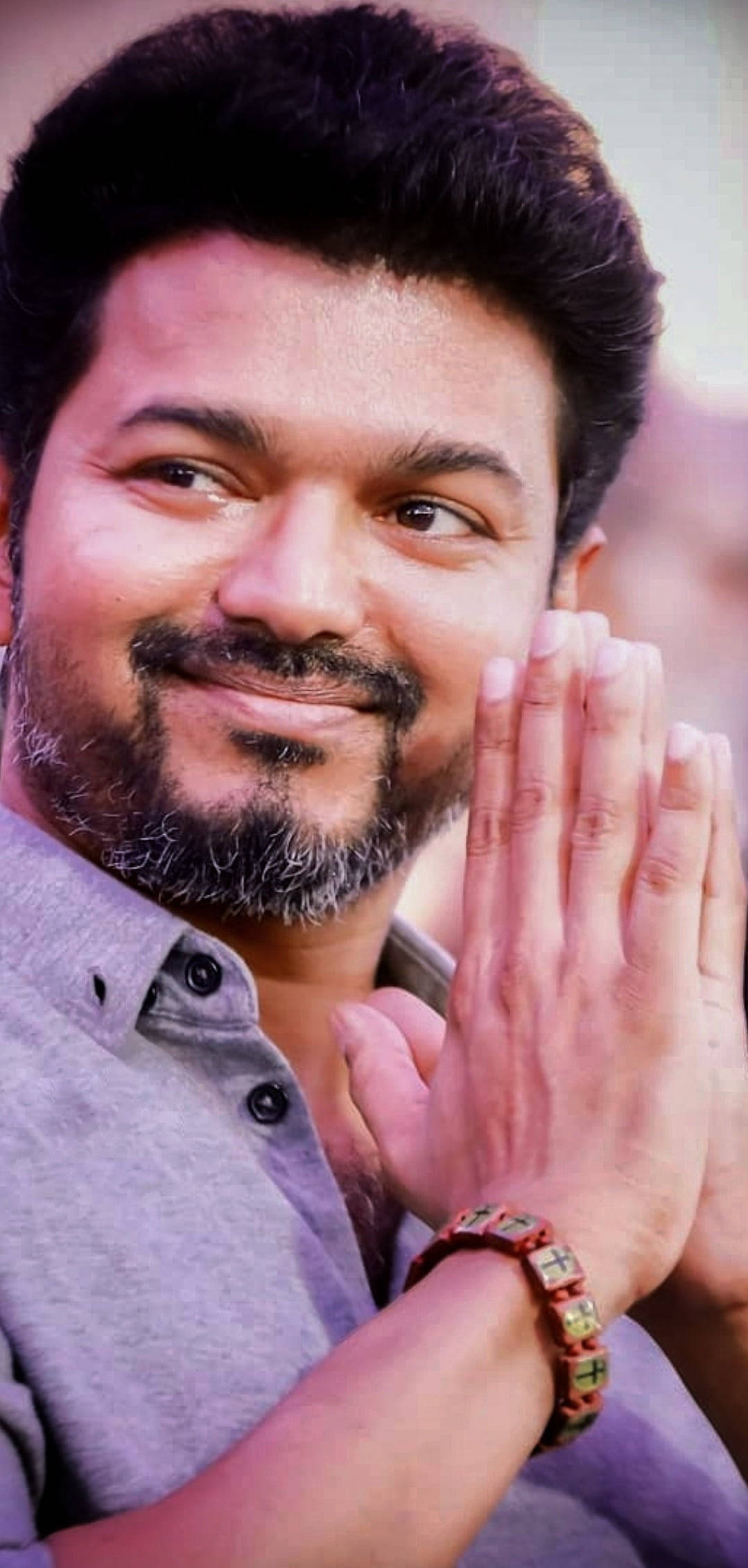 vijay HD 桌布 壁紙 — 一位面帶溫暖笑容、留鬍鬚的男子，雙手合十，恭敬地做手勢，身穿灰色襯衫和珠子手鍊。
