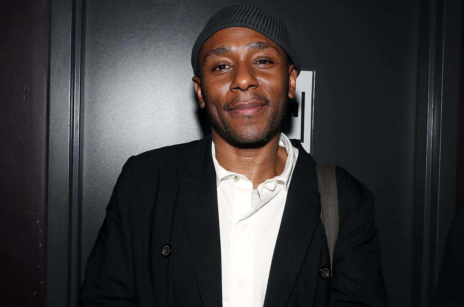 Yasiin Bey 桌布 壁紙 — 一位面帶溫暖笑容的男子，在深色背景前穿著黑色毛帽和西裝外套，內搭白色襯衫。
