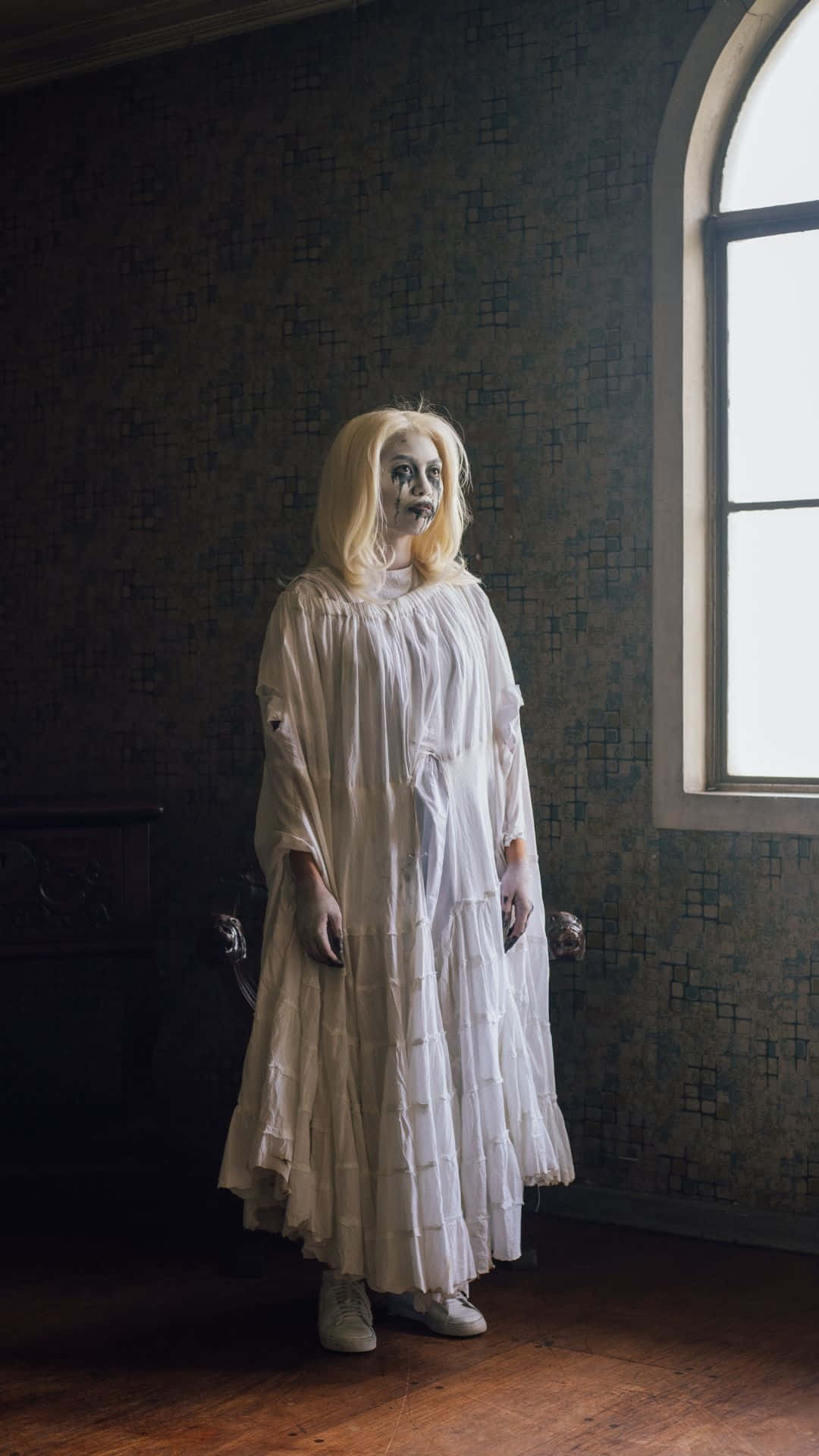 Horror Costumes 桌布 壁紙 — 一位面色蒼白的女人穿著破爛的白裙，詭異地站在一個光線昏暗、有圖案壁紙和拱形窗戶的房間裡。