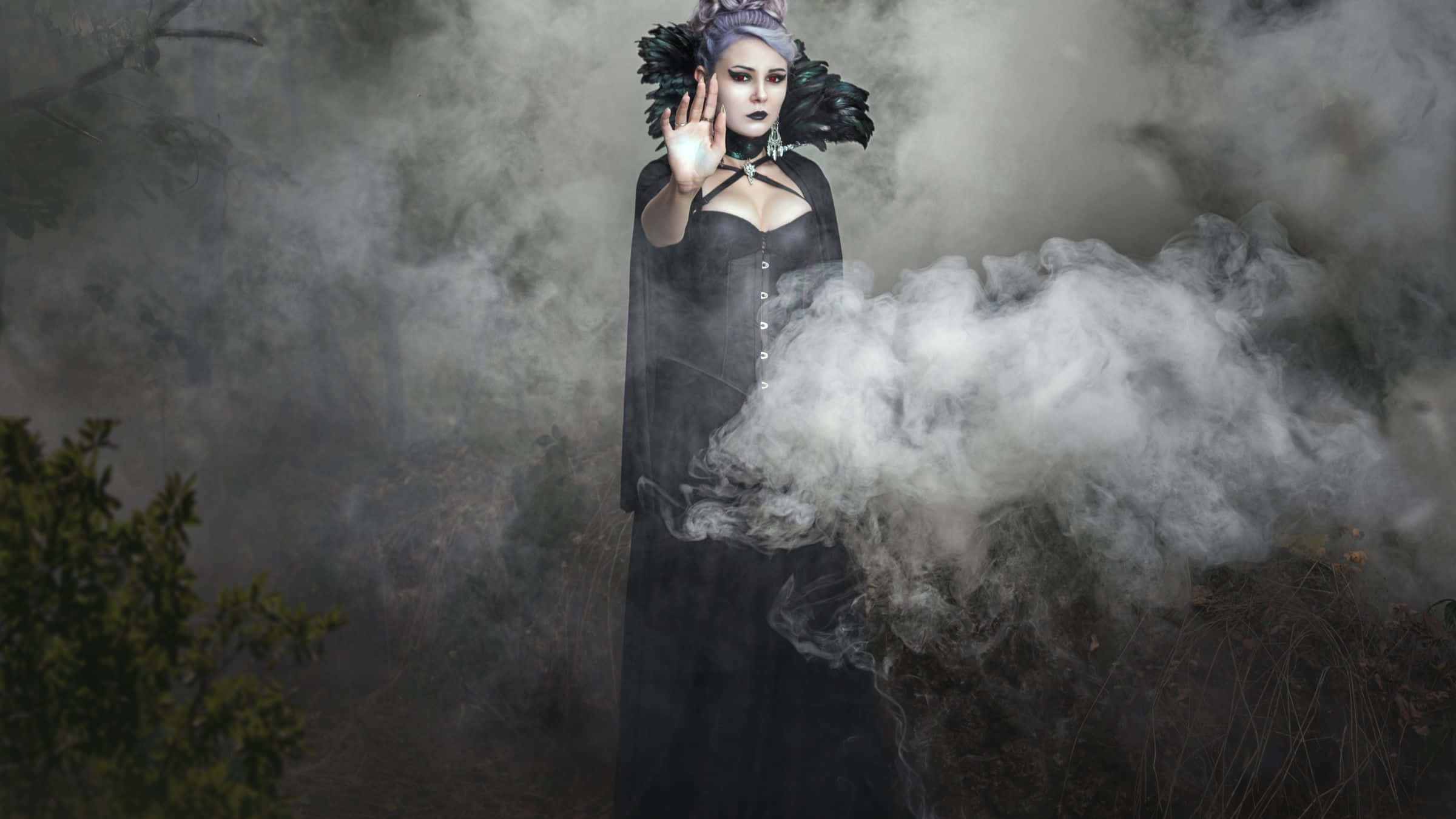 Goth 桌布 壁紙 — 一位面色蒼白的女性，身穿黑色長袍和羽毛頭飾，站在迷霧森林中，舉起手做出停止手勢。