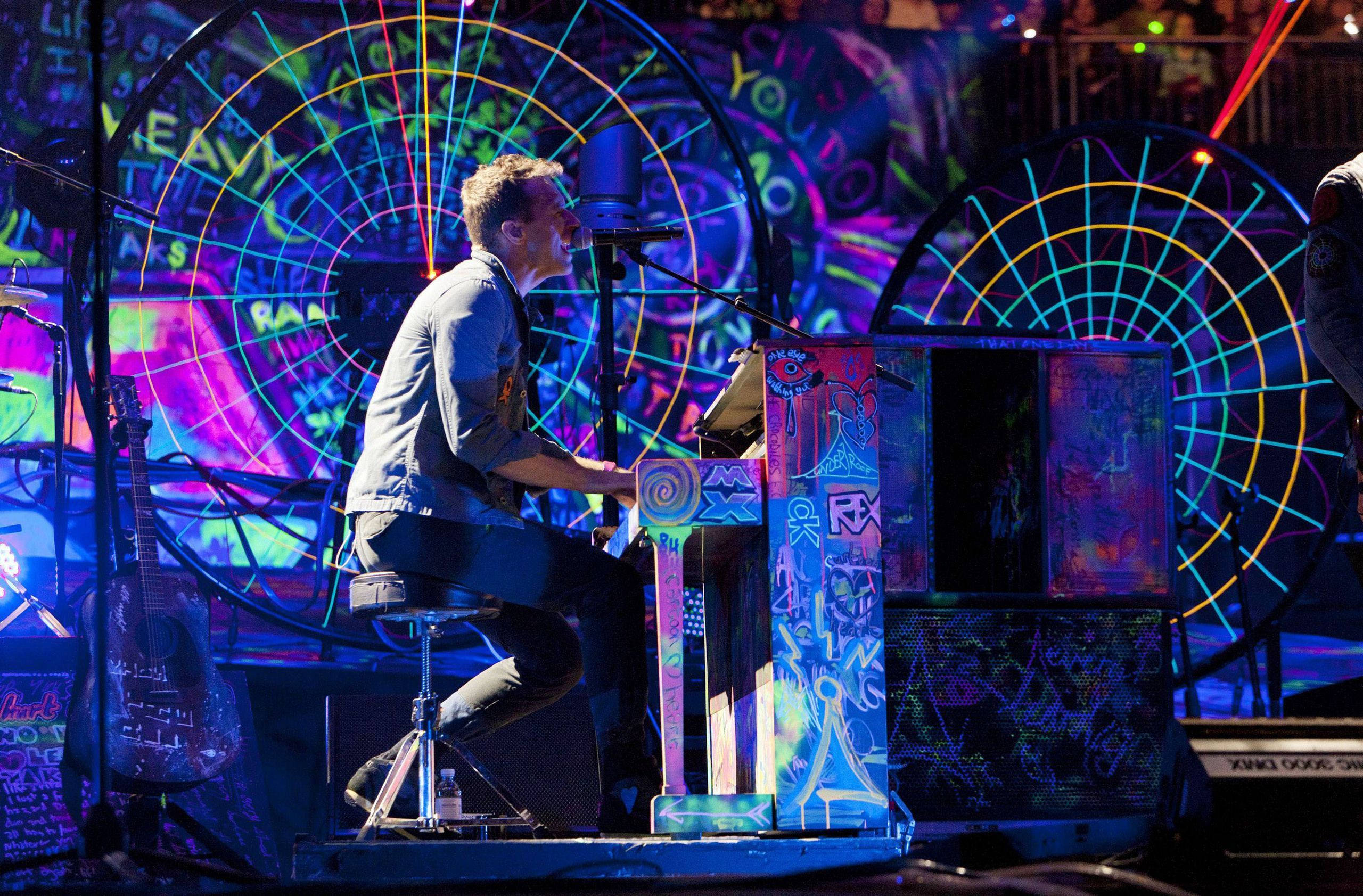 Chris Martin 桌布 壁紙 — 一位音樂家在充滿活力的舞台上演奏塗鴉鋼琴，舞台以霓虹燈和圓形投影照亮。