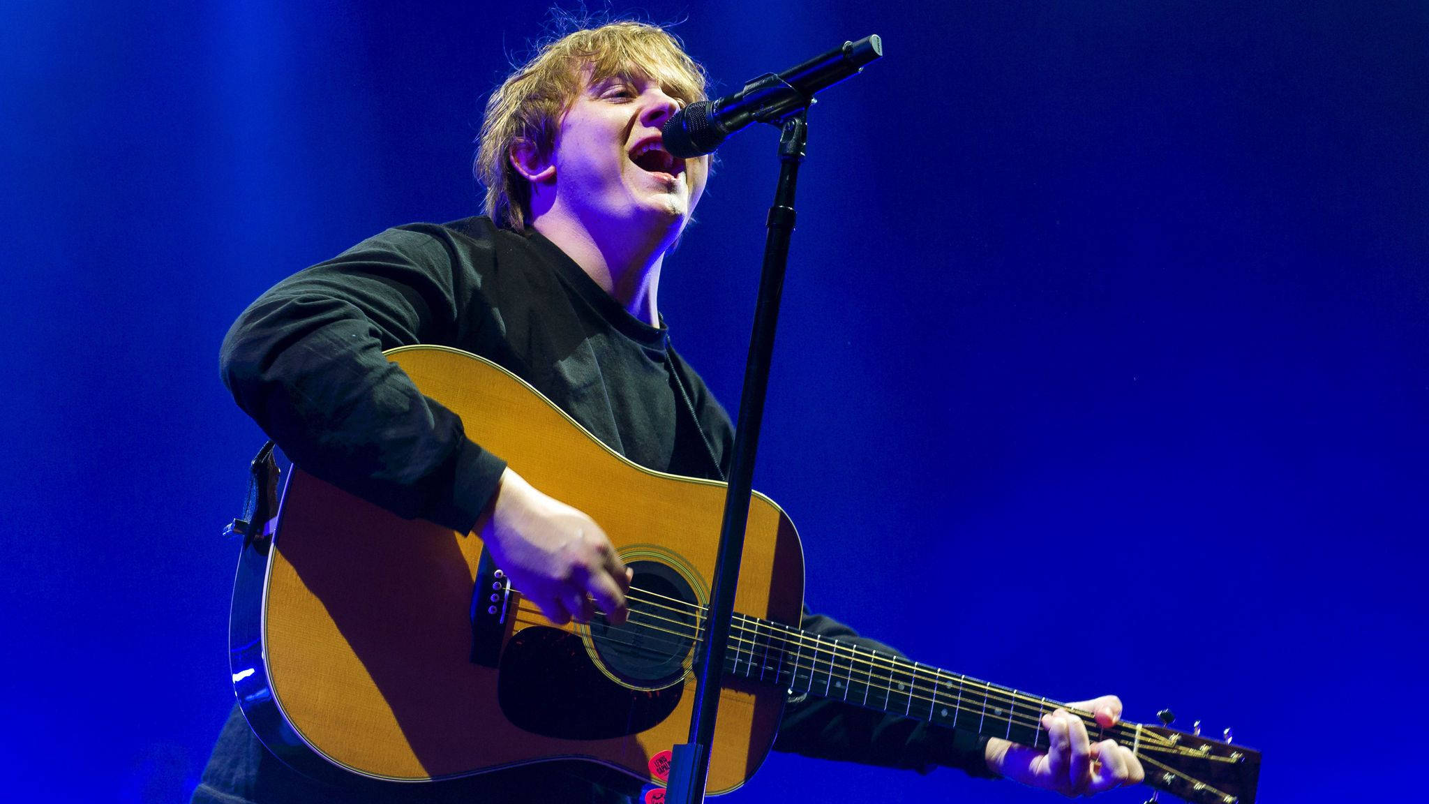 Lewis Capaldi 桌布 壁紙 — 一位音樂家在充滿活力的藍色舞台燈光下，一邊彈奏木吉他，一邊熱情地對著麥克風唱歌。
