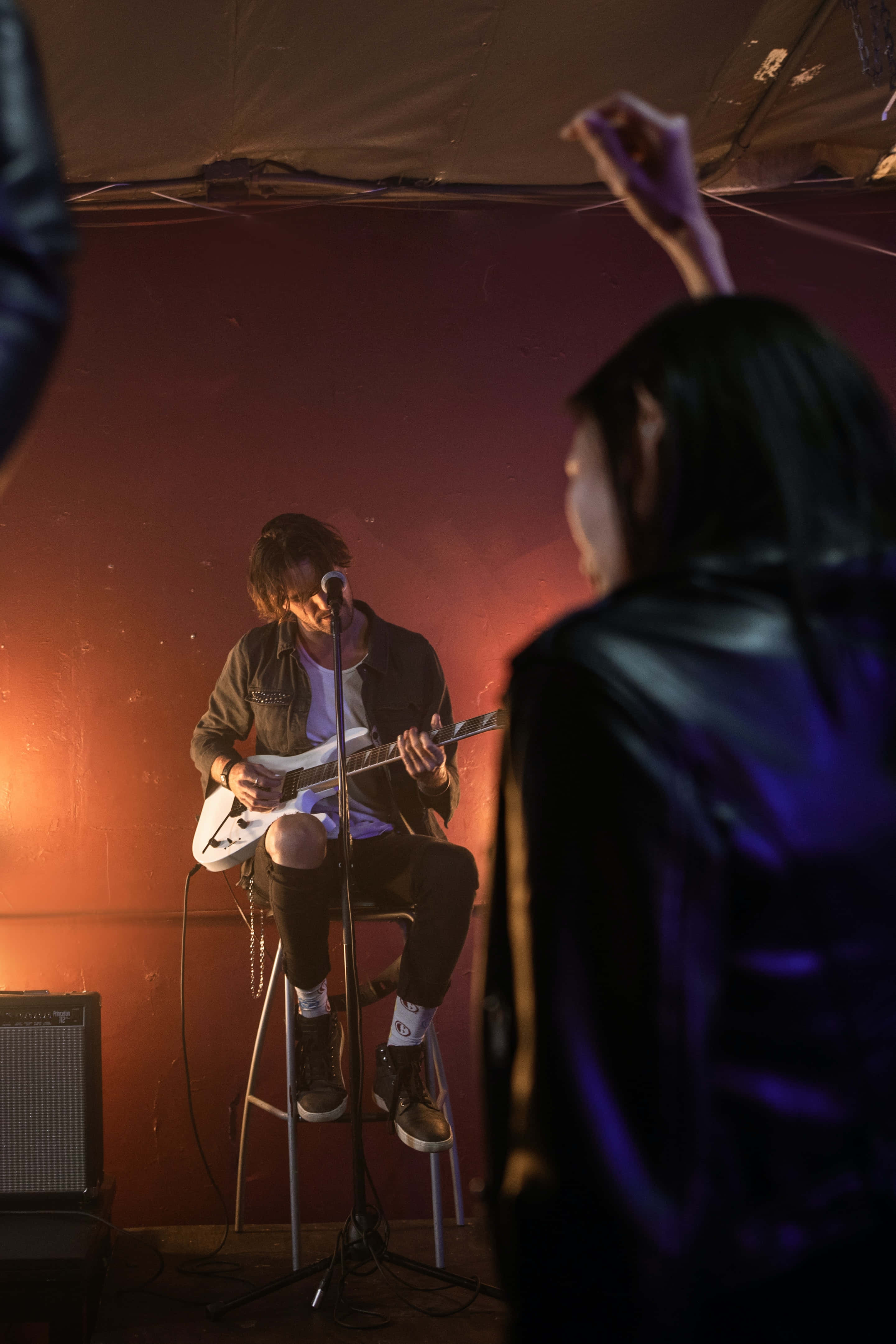Singer 桌布 壁紙 — 一位音樂家在溫暖的舞台燈光下坐在凳子上彈奏電吉他，而一位觀眾在紅牆前舉起手。