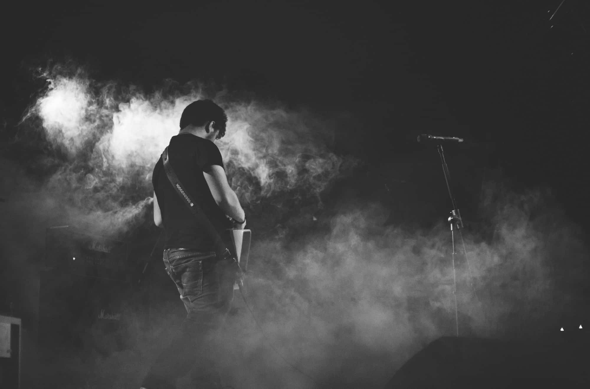 Singer 桌布 壁紙 — 一位音樂家在煙霧瀰漫的舞台上，在戲劇性燈光下彈吉他的黑白演唱會照片。