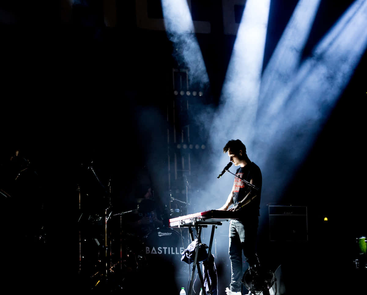 Bastille 桌布 壁紙 — 一位音樂家在煙霧瀰漫的音樂會舞台上，在戲劇性的藍色聚光燈下彈奏鍵盤。