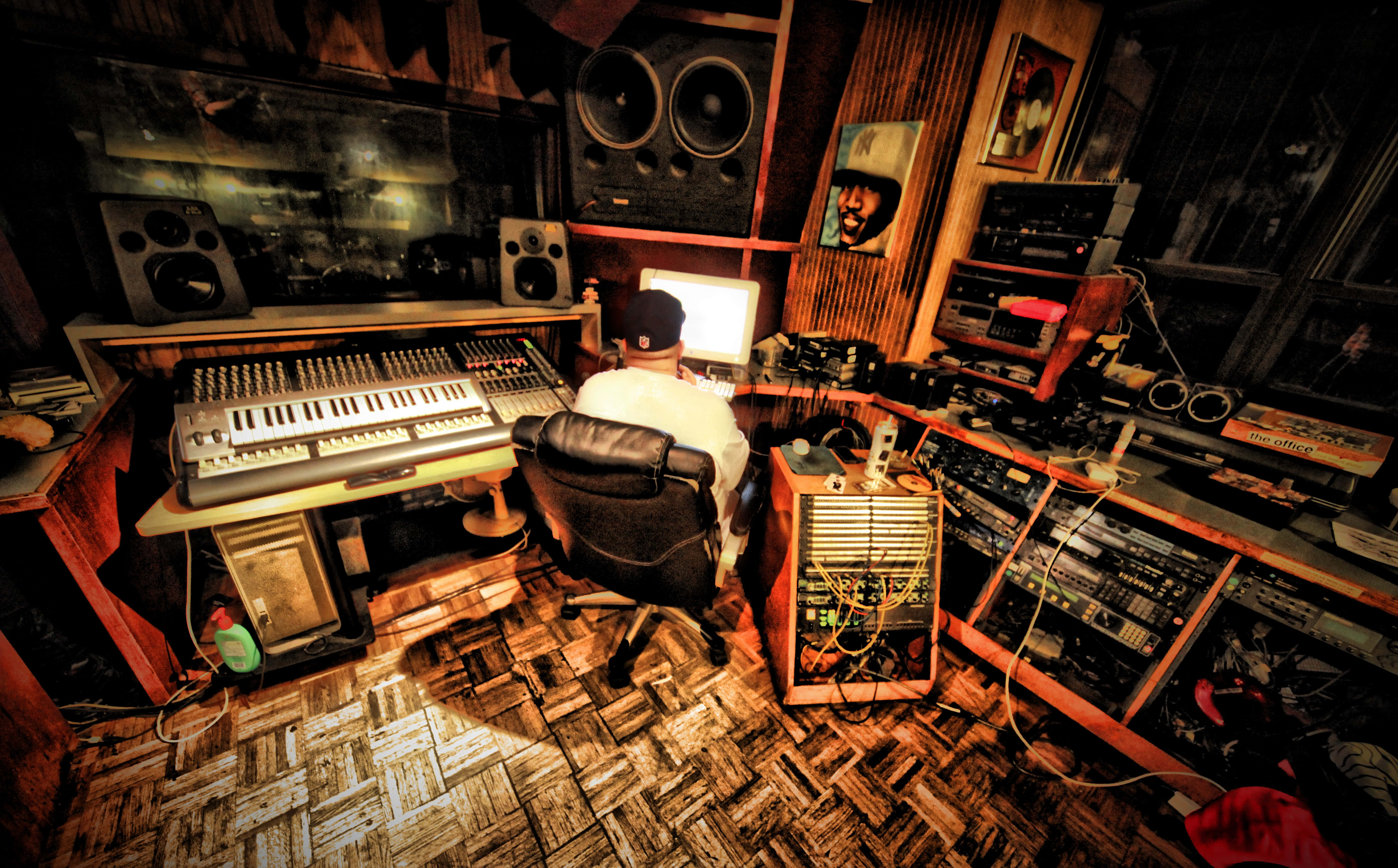 Fl Studio 桌布 壁紙 — 一位音樂家坐在雜亂的工作室桌前，周圍環繞著復古音響設備和發光的螢幕，在溫暖的琥珀色燈光下。