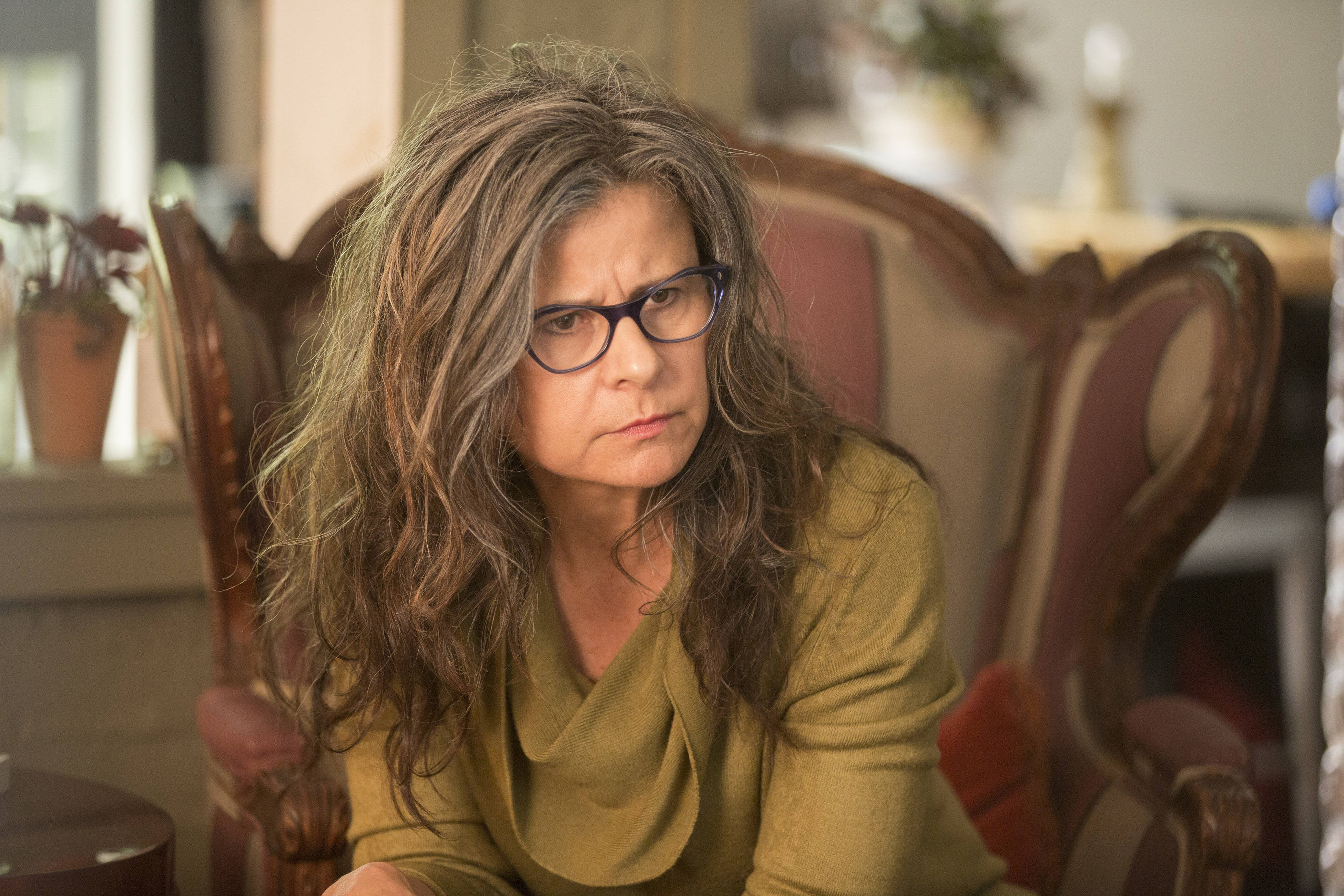 Tracey Ullman 桌布 壁紙 — 一位頭髮凌亂灰白、戴著眼鏡的女性，坐在一張復古椅子上，在溫暖的燈光下顯得很惱怒。