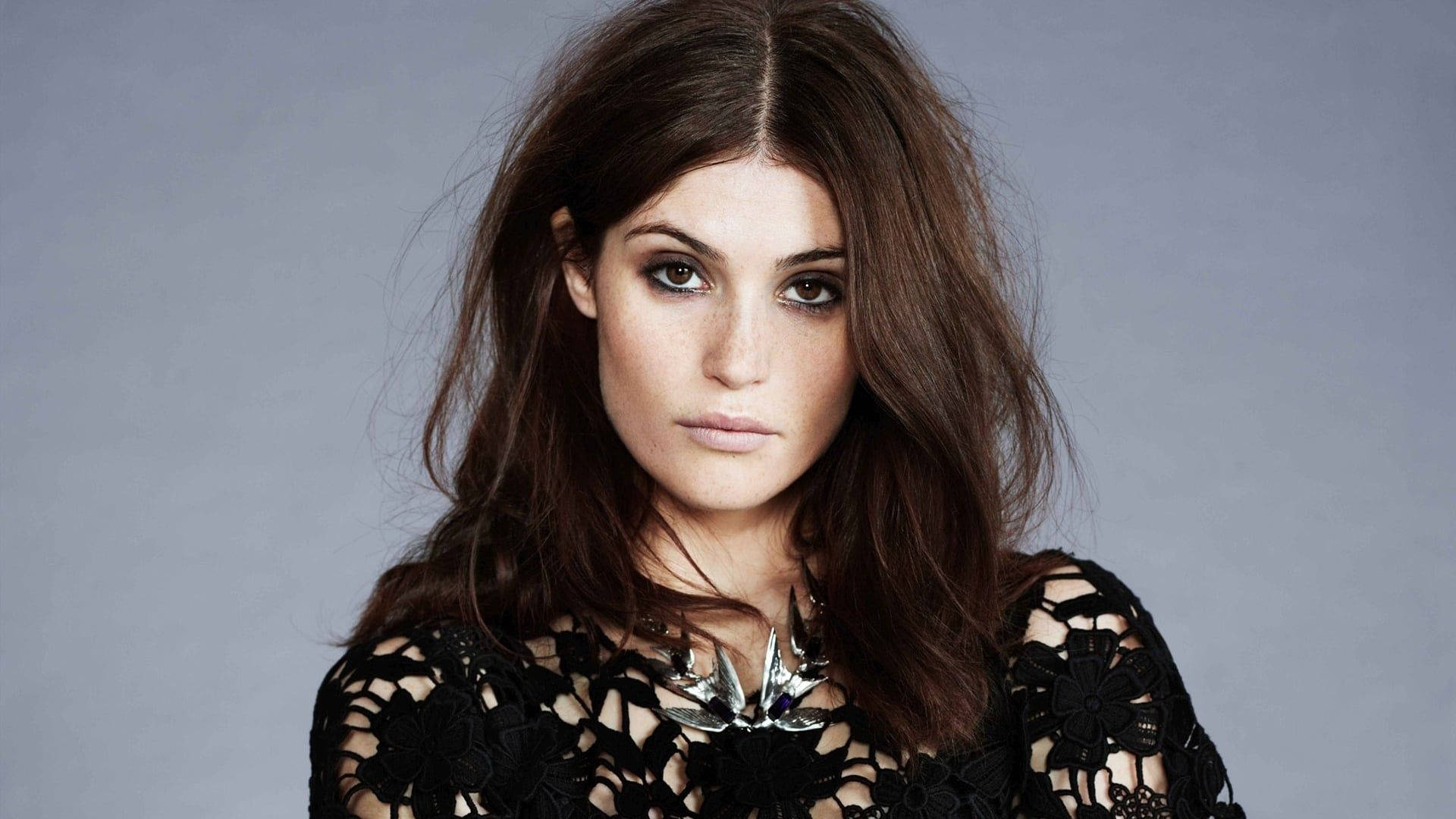 Gemma Arterton 桌布 壁紙 — 一位頭髮凌亂的棕色頭髮、眼神強烈的女性，在柔和的灰色背景前穿著黑色蕾絲上衣。