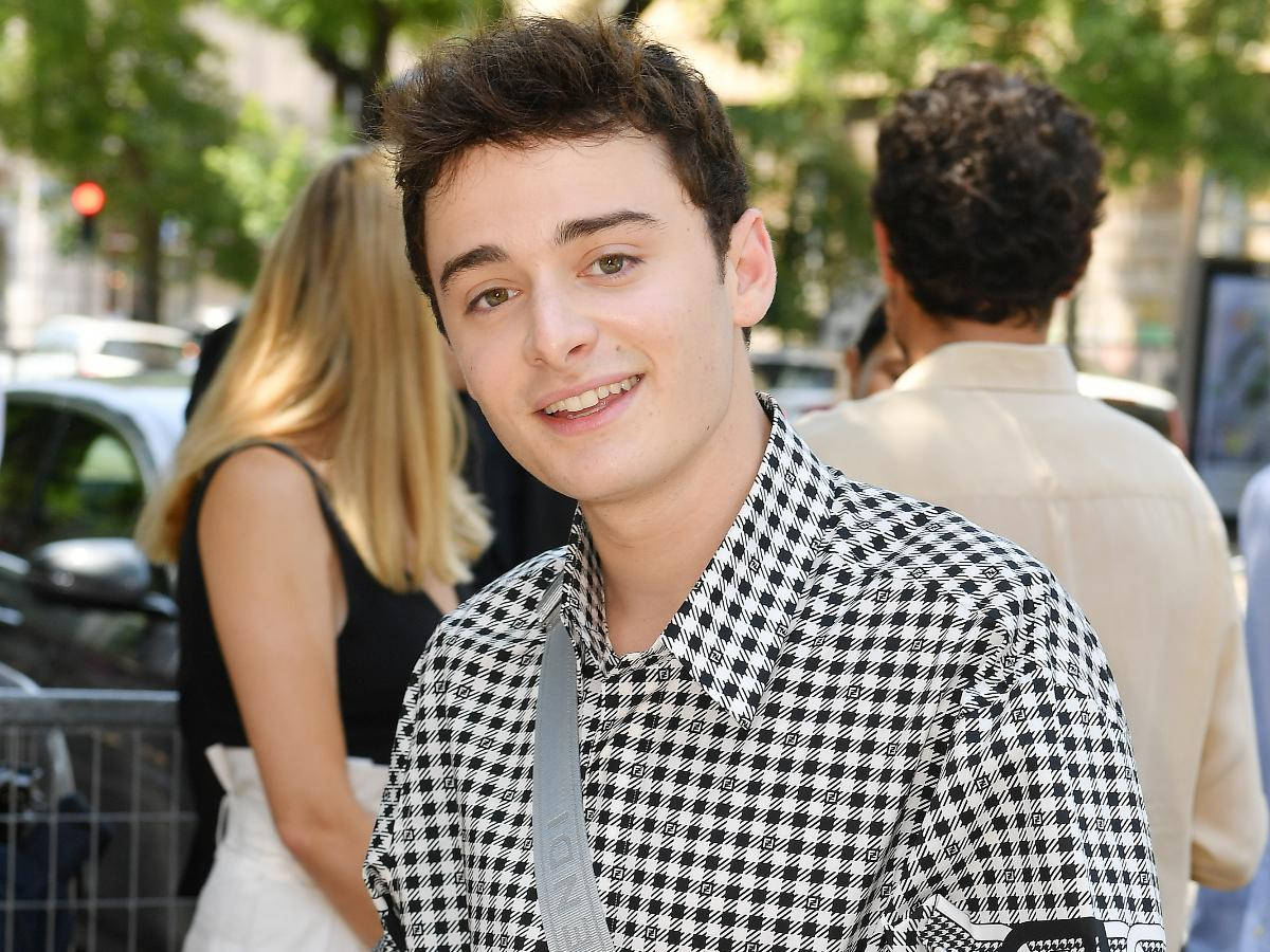 Celebrity 桌布 壁紙 — 一位頭髮凌亂的深色頭髮年輕男子，在陽光明媚的城市街道上穿著黑白千鳥格襯衫，露出溫暖的笑容。