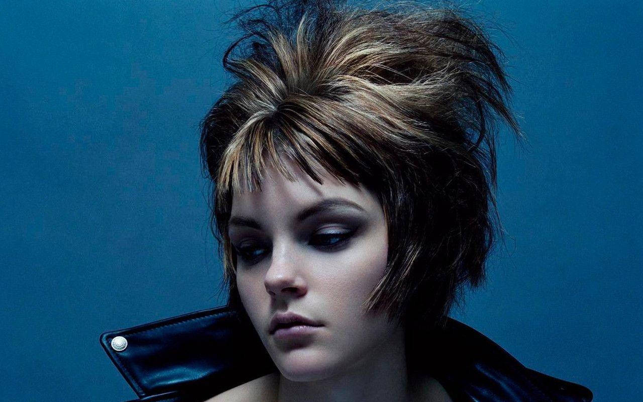 jessica stam 桌布 壁紙 — 一位頭髮尖刺蓬亂、化著深色妝容的女性，穿著黑色皮夾克，在純藍色背景前向下凝視。