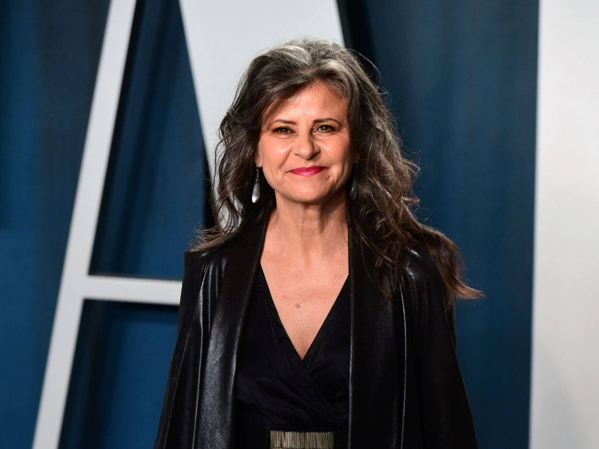 Tracey Ullman 桌布 壁紙 — 一位頭髮帶有灰色挑染波浪捲的女性，身穿黑色皮夾克，在藍白幾何背景前溫暖微笑。
