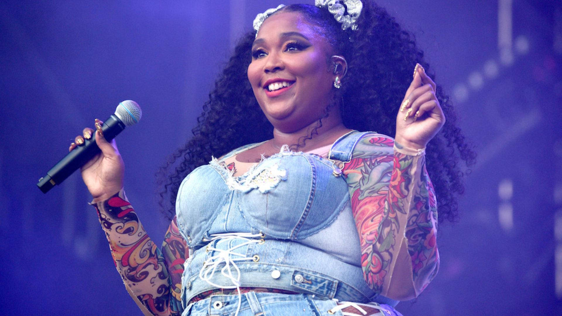 Lizzo 桌布 壁紙 — 一位頭髮捲曲、帶有彩色刺青的快樂歌手在紫色燈光下拿著麥克風在舞台上表演。