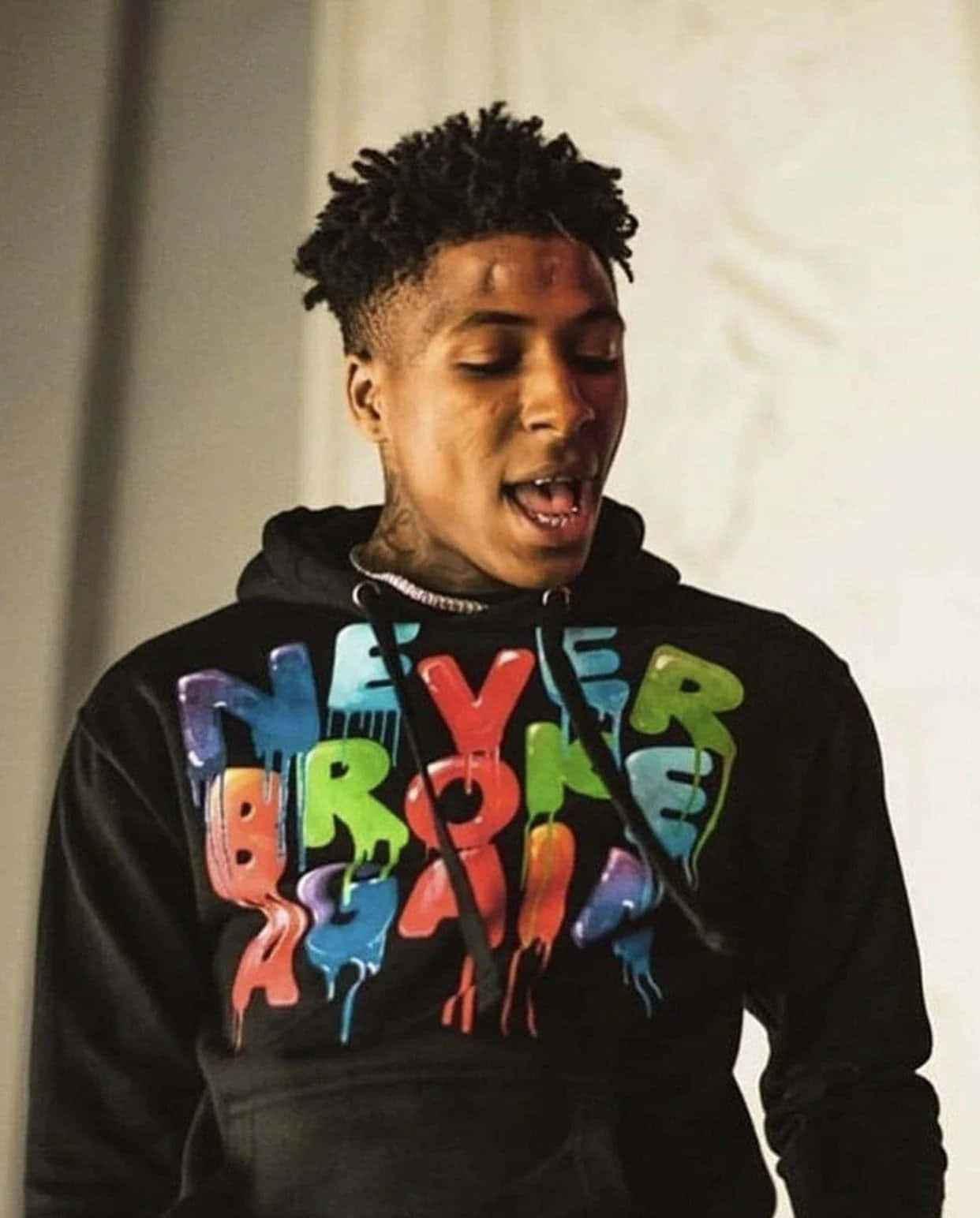 NBA YoungBoy 桌布 壁紙 — 一位頭髮有紋理的年輕男子穿著黑色連帽衫，上面有鮮豔滴落的彩虹文字寫著「NEVER BROKE AGAIN」，正在唱歌。