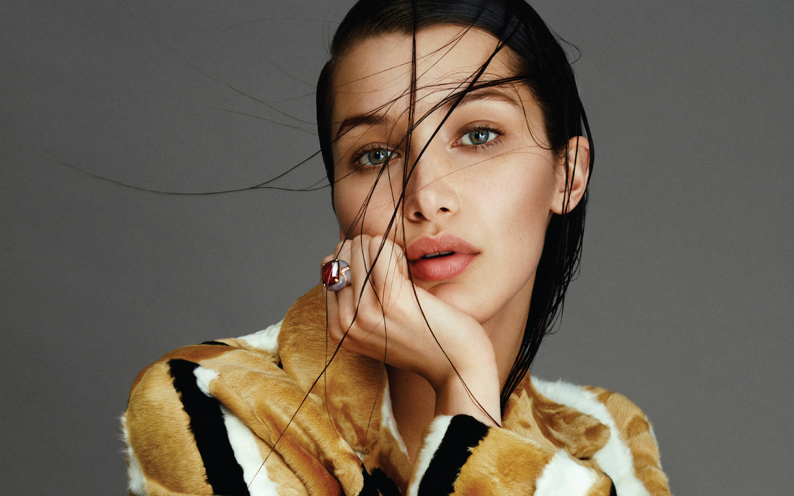 Bella Hadid 桌布 壁紙 — 一位頭髮濕透、擁有引人注目的綠色眼睛的女性，將下巴擱在手上，直視鏡頭，身穿虎紋毛皮大衣，背景為中性灰色。