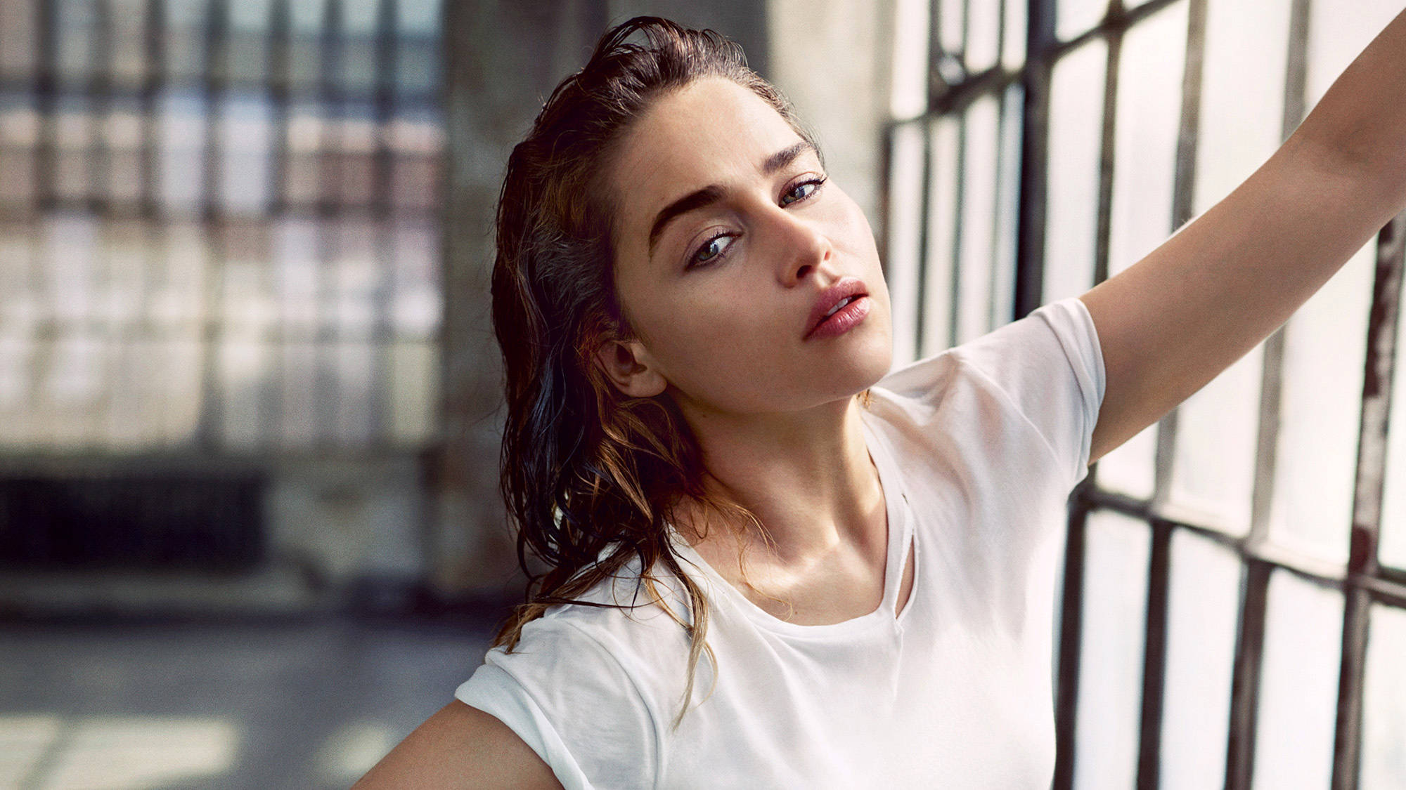 Emilia 桌布 壁紙 — 一位頭髮濕透、身穿白色T恤的女性在陽光普照的城市閣樓的大窗戶附近伸展身體。