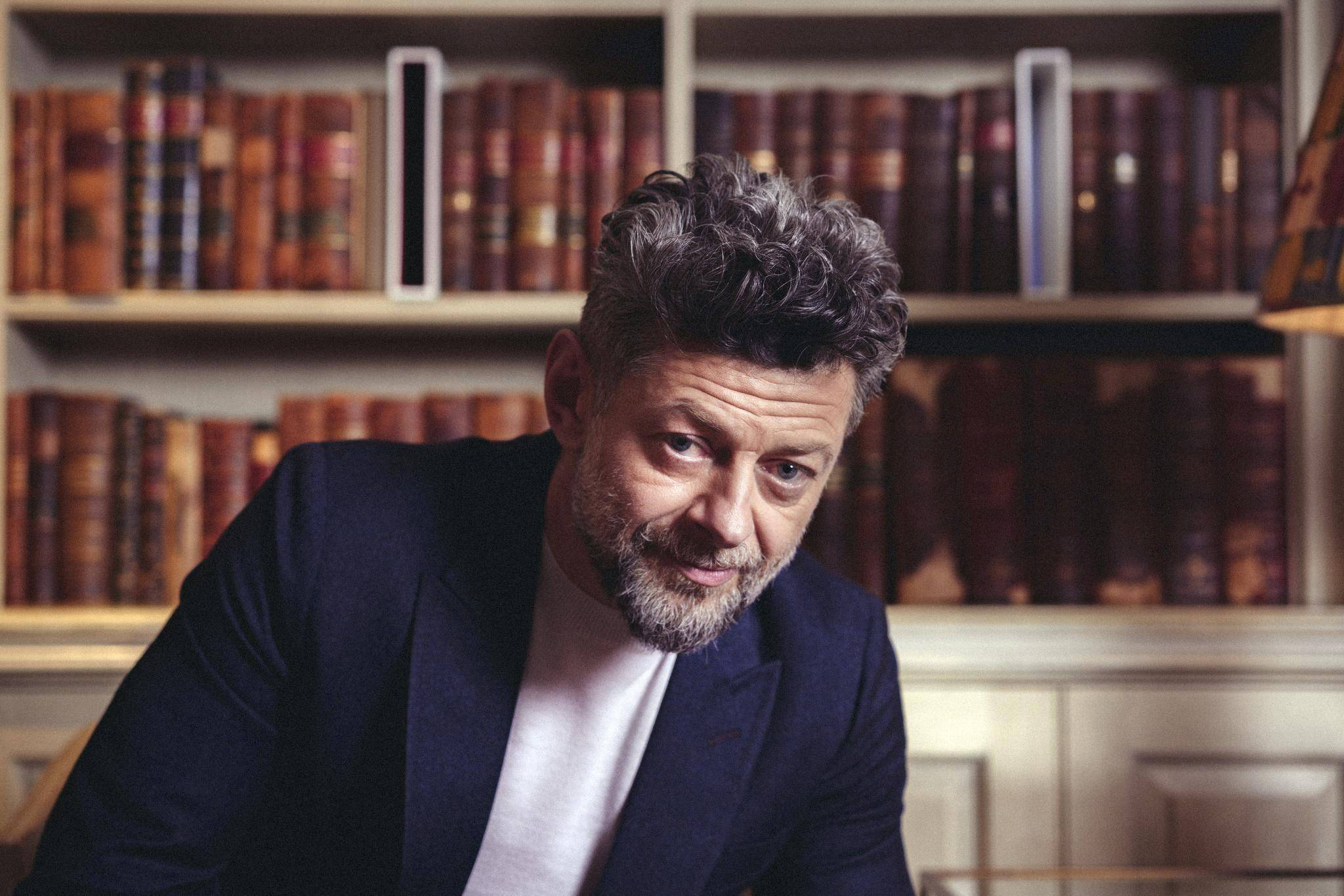 actor 桌布 壁紙 — 一位頭髮花白、留著鬍鬚的男子，身穿深色西裝外套，在溫暖燈光的圖書館裡向前傾，身後是木質書架。