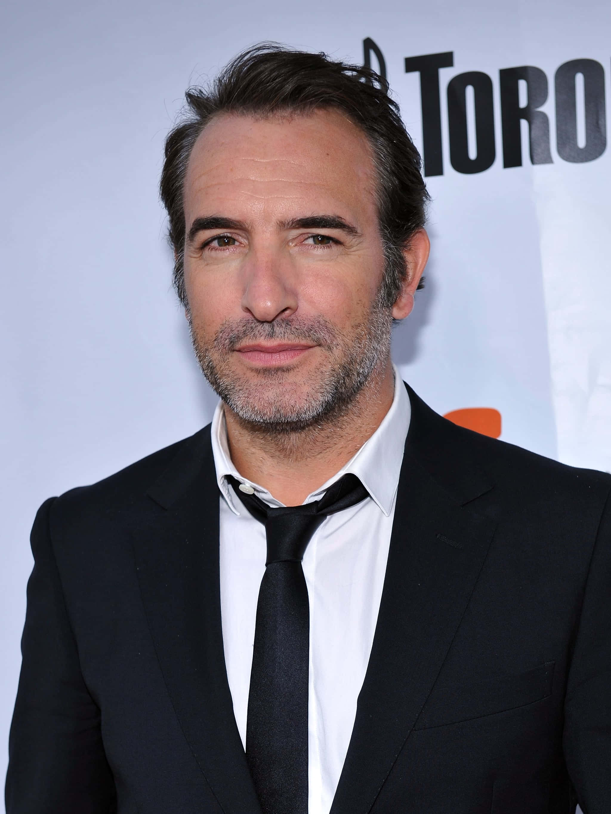 Jean Dujardin 桌布 壁紙 — 一位頭髮花白、留鬍鬚的男子，身穿黑色西裝打領帶，背景為白色並帶有「TORO」文字。