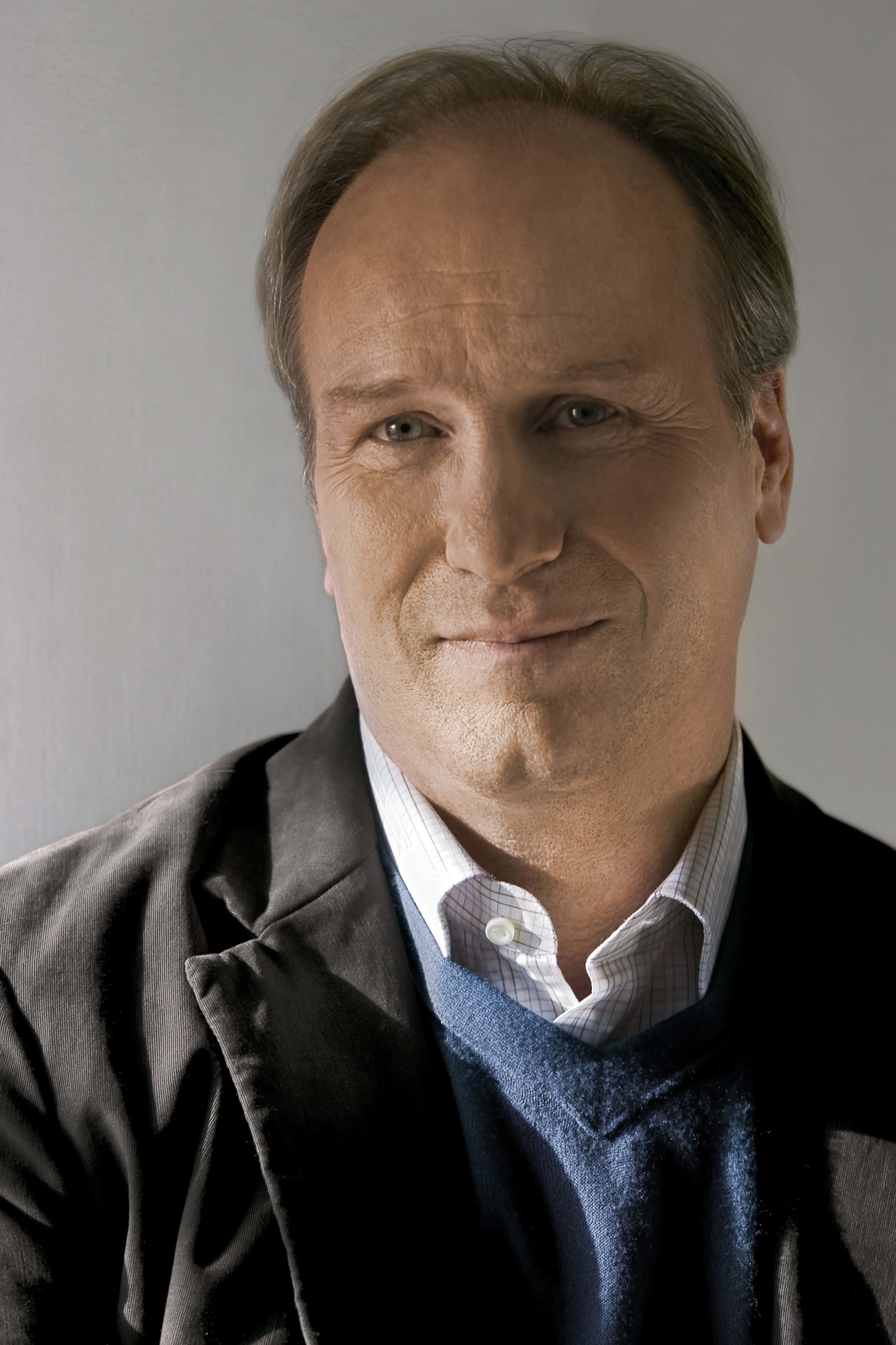 William Hurt 桌布 壁紙 — 一位頭髮花白的中年男子，身穿深色西裝外套，內搭藍色毛衣和格子襯衫，在純灰色背景前溫柔微笑。
