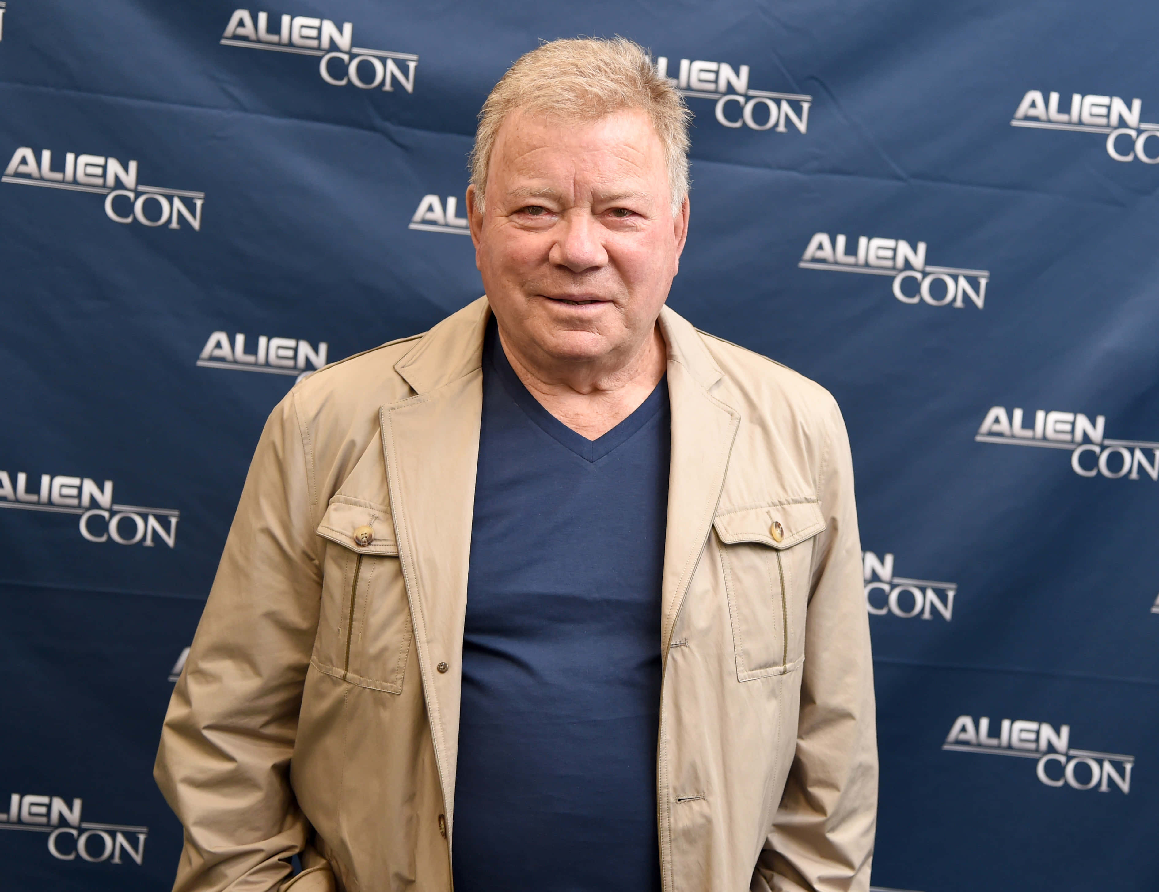 william shatner 桌布 壁紙 — 一位頭髮花白的男士穿著米色外套和藍色襯衫，在重複的外星人展背景前對著鏡頭微笑。