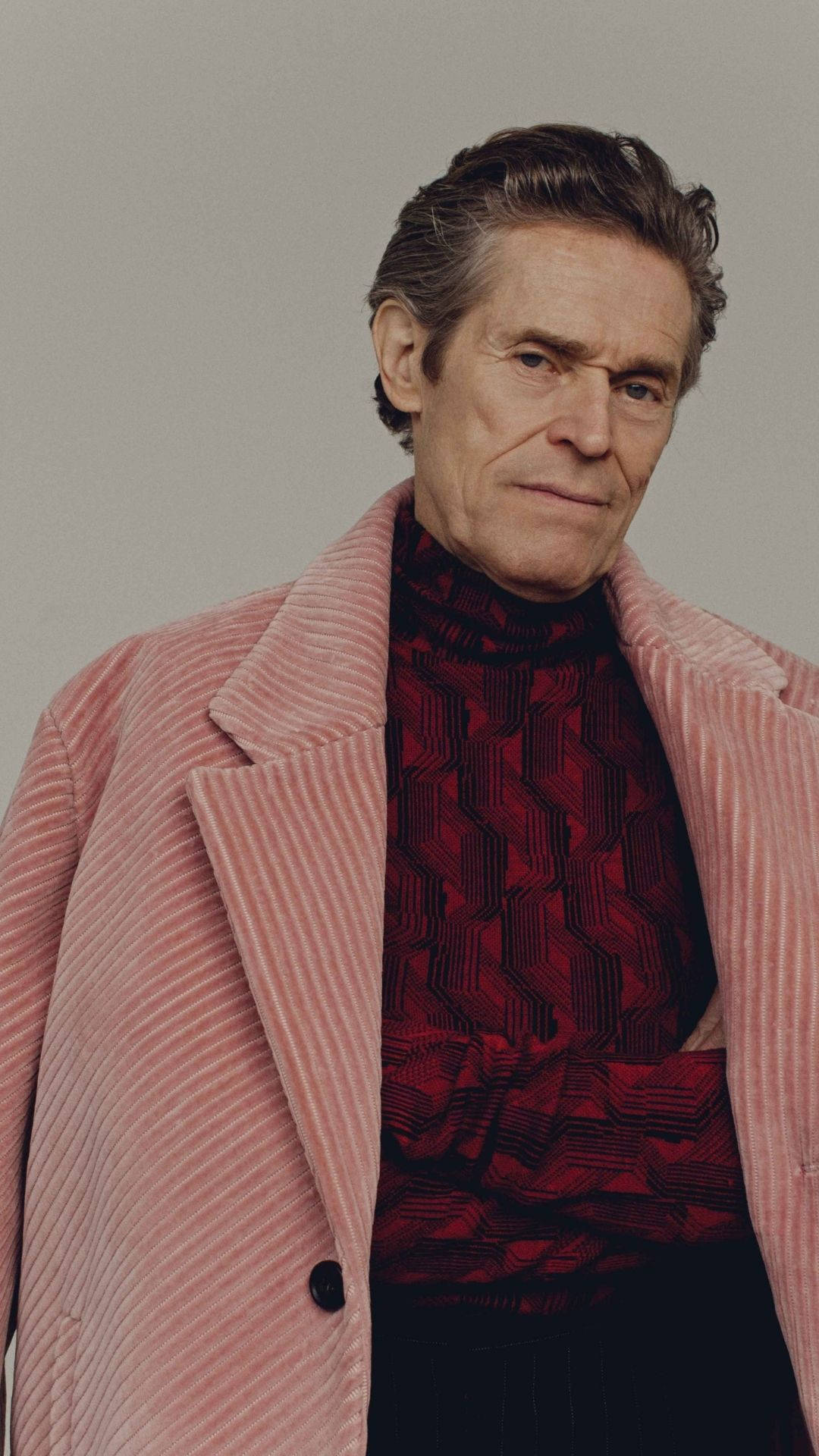 Willem Dafoe 桌布 壁紙 — 一位頭髮花白的男士穿著粉紅色燈芯絨外套，內搭紅色幾何圖案高領毛衣，背景為中性灰色。