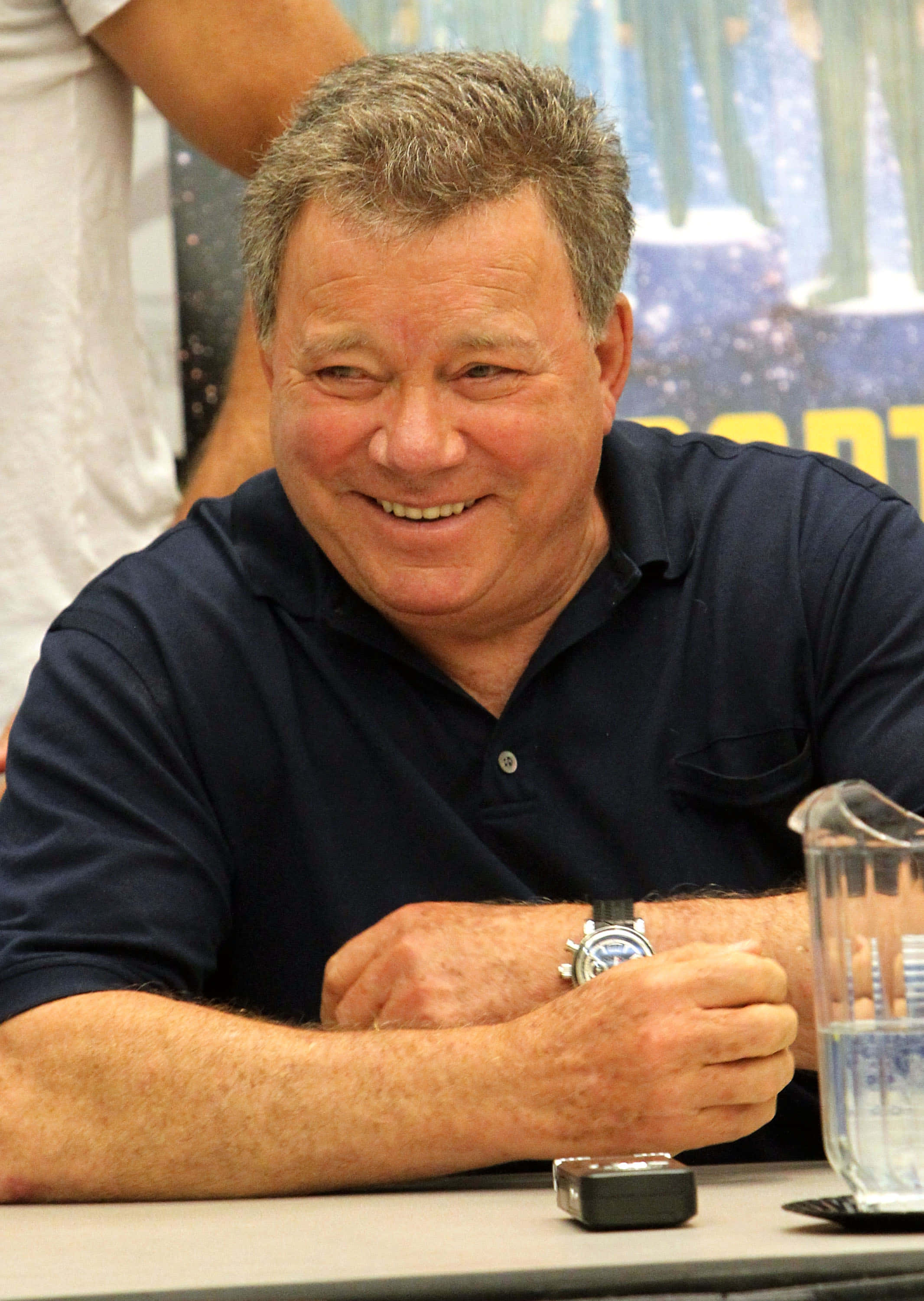 william shatner 桌布 壁紙 — 一位頭髮花白的開朗男士，身穿深藍色Polo衫，坐在桌前露出溫暖的笑容。