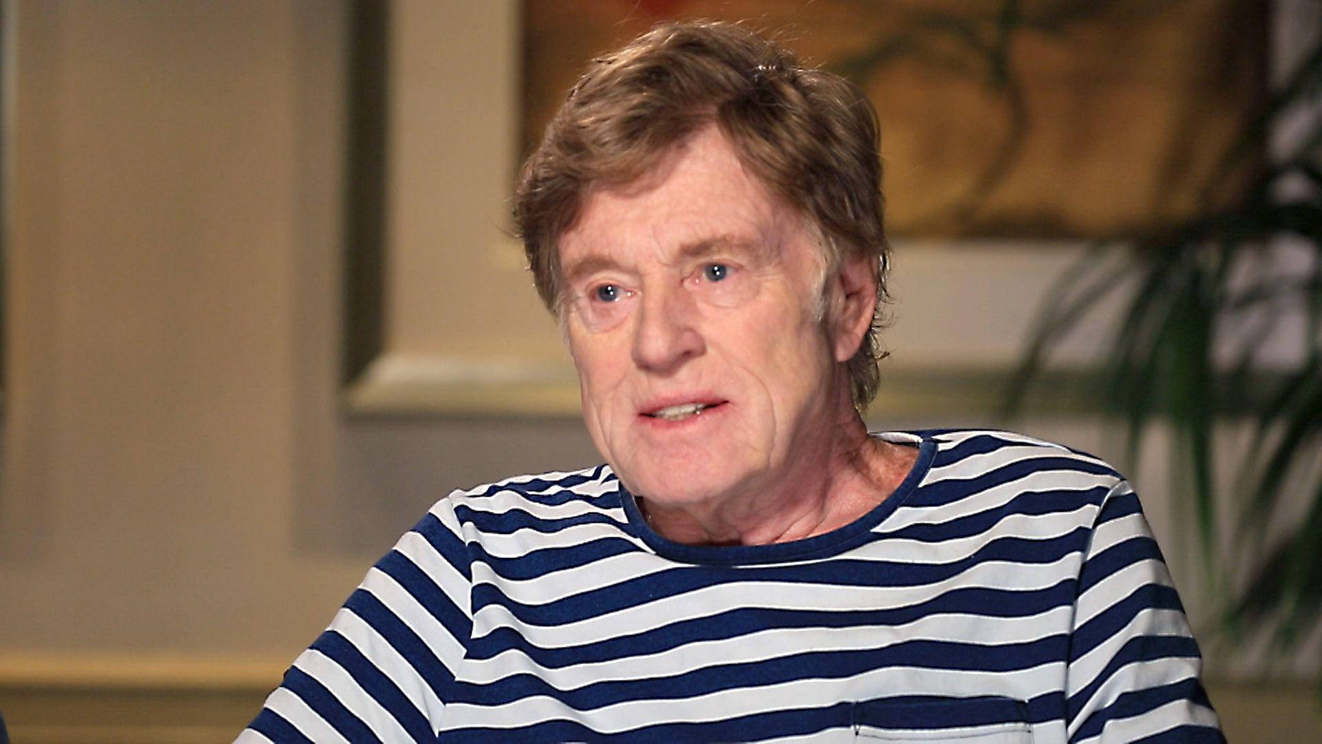 Celebrity 桌布 壁紙 — 一位頭髮蓬亂的年長男士穿著藍白條紋襯衫，坐在室內柔和燈光下。