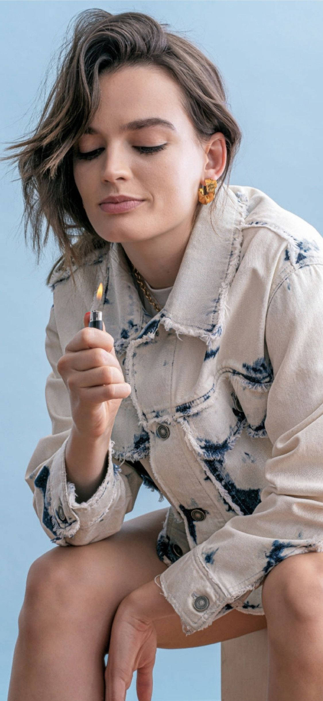 Emma 桌布 壁紙 — 一位頭髮蓬亂的棕髮女性，平靜地坐在柔和的藍色背景前，手持點燃的打火機。