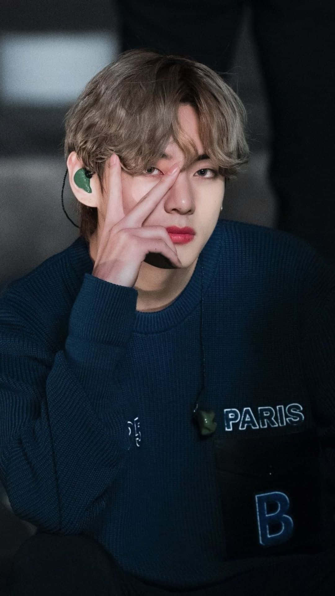 BTS 桌布 壁紙 — 一位頭髮蓬鬆、嘴唇紅潤的年輕男子，身穿繡有「PARIS」和「B」的深藍色毛衣，將手指放在眼睛上擺姿勢。