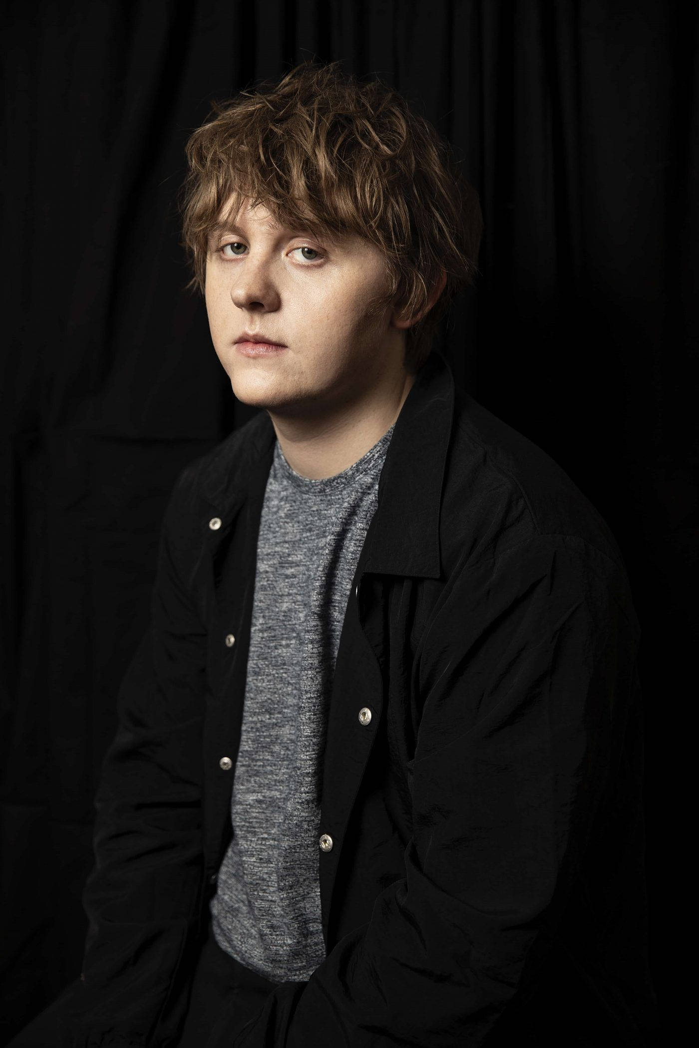 Lewis Capaldi 桌布 壁紙 — 一位頭髮蓬鬆的年輕男子在鮮明的黑色背景下，若有所思地凝視著，營造出充滿氛圍的攝影棚肖像。