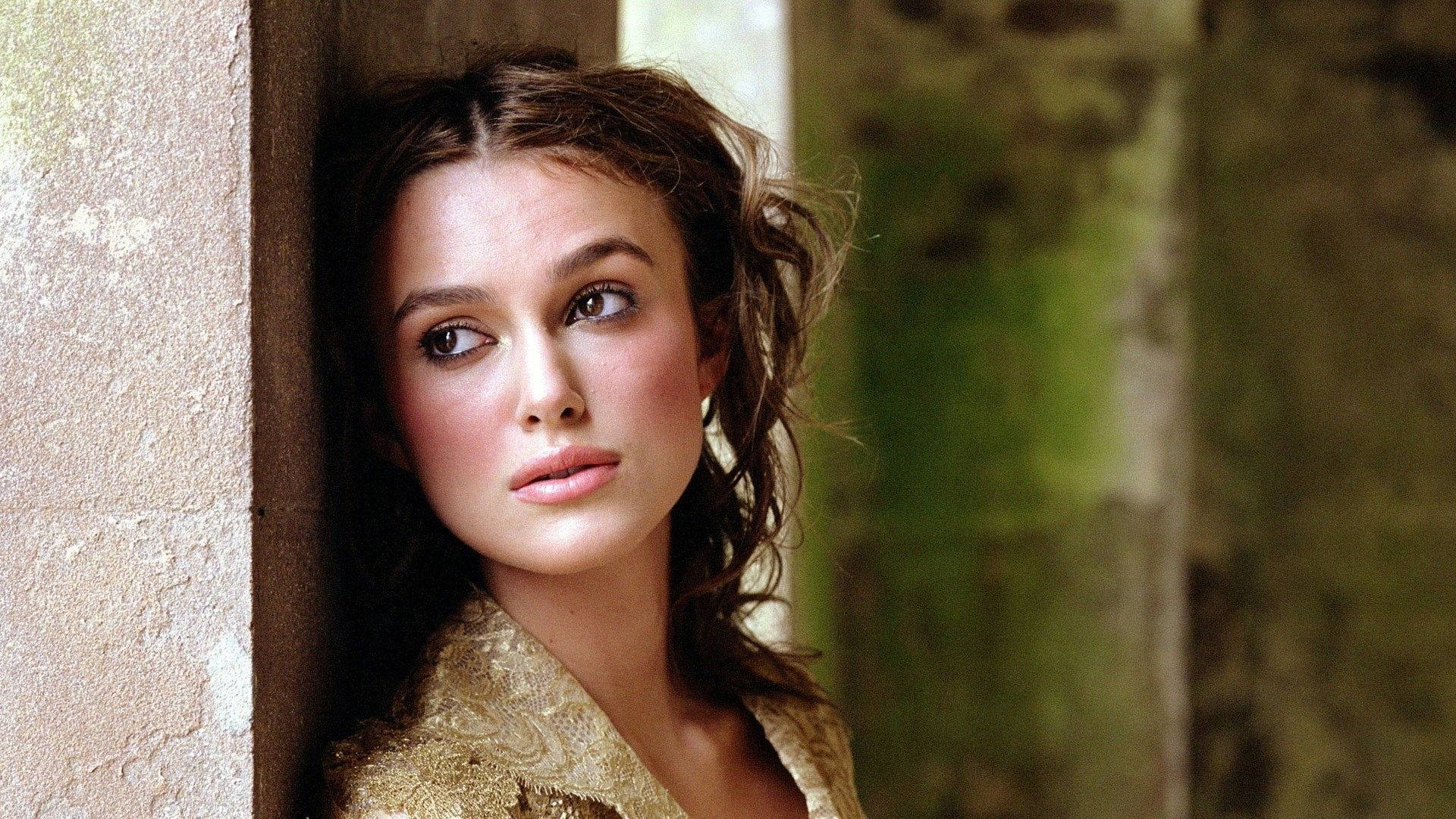 Keira Knightley 桌布 壁紙 — 一位頭髮蓬鬆的棕色頭髮女士靠在石牆上，在柔和自然光線中若有所思地凝視遠方。