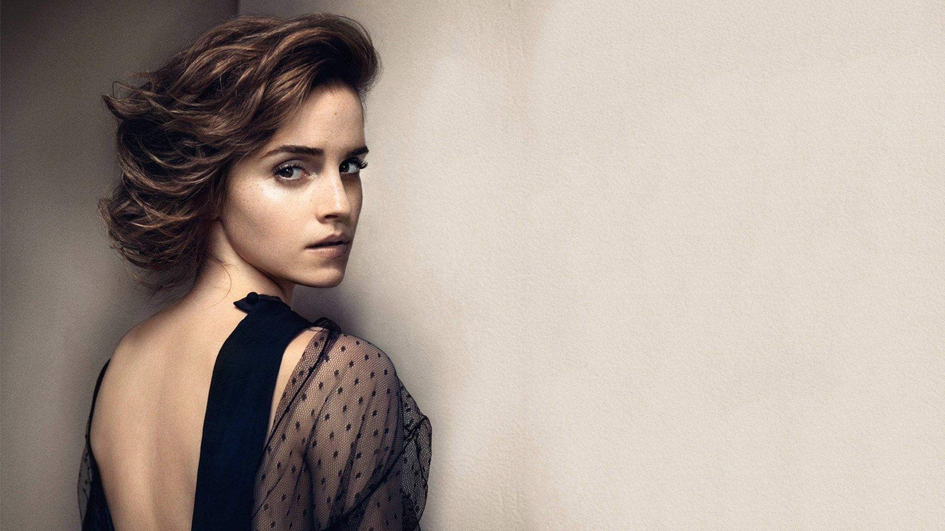 Emma 桌布 壁紙 — 一位頭髮蓬鬆的棕色頭髮女性靠著素色牆壁，身穿黑色透視波點上衣。