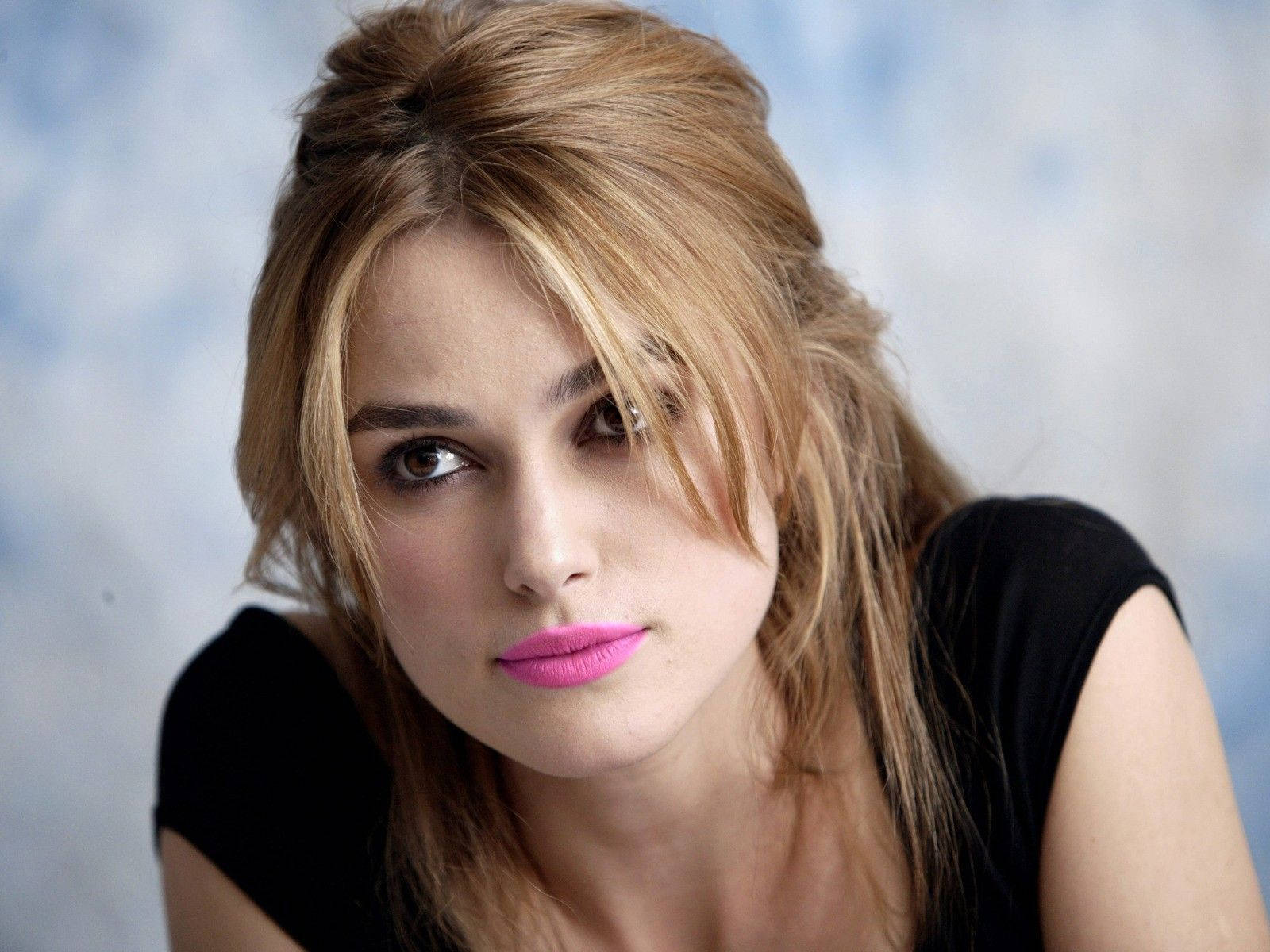 Keira Knightley 桌布 壁紙 — 一位頭髮蓬鬆的金髮女子，塗著大膽的粉紅色唇膏，在柔和的藍色紋理背景下凝視著。