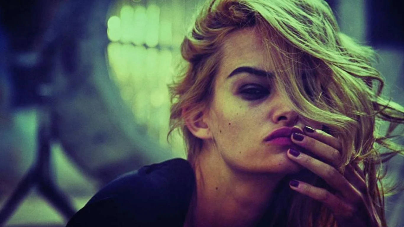 attitude 4K 桌布 壁紙 — 一位頭髮蓬鬆的金髮女子塗著深色唇膏，手遮住部分臉龐，在模糊的綠色背景下若有所思地凝視著。