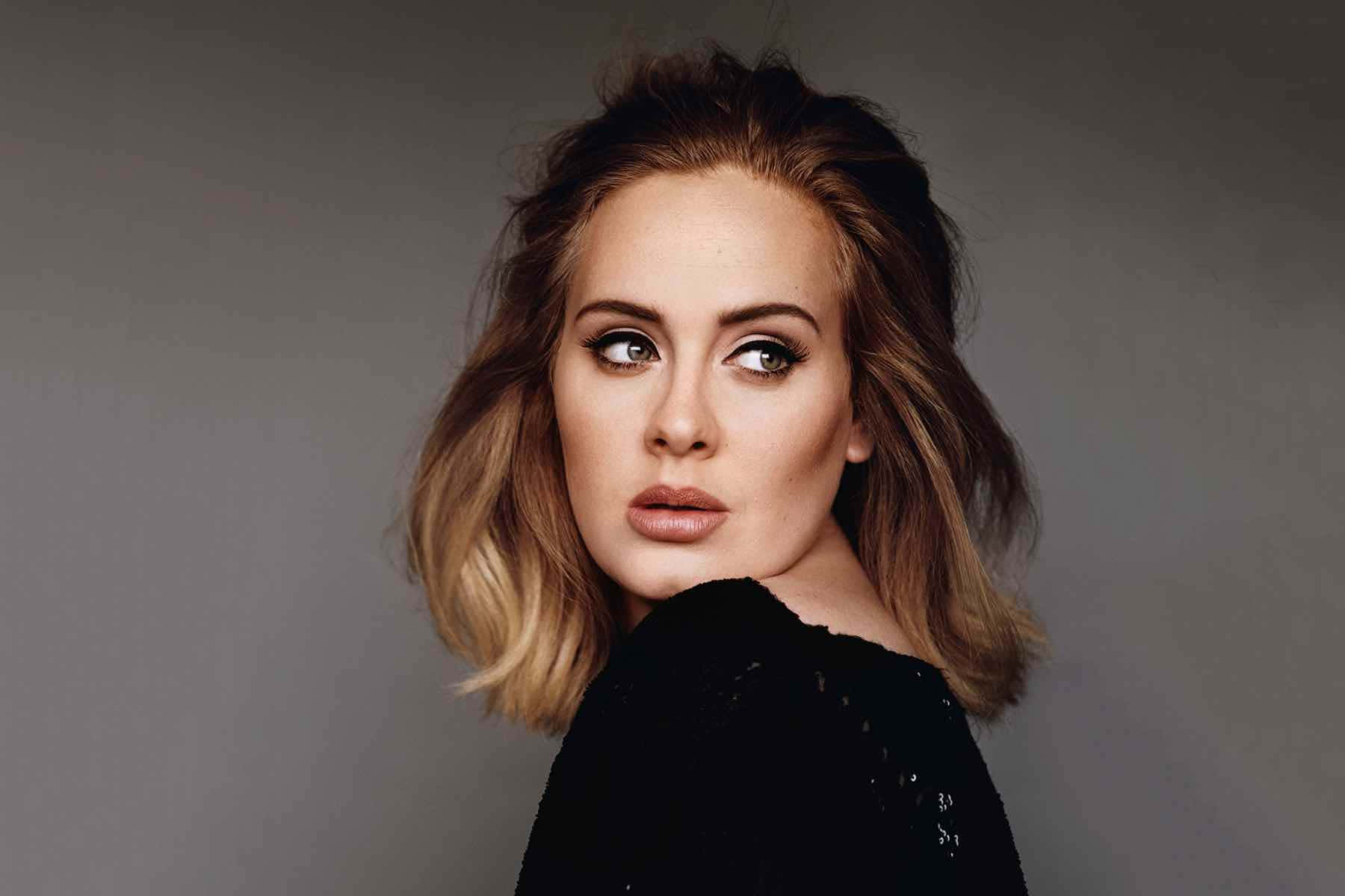 Adele 桌布 壁紙 — 一位頭髮蓬鬆金髮、妝容大膽的女性，在柔和的灰色背景下側視，身穿黑色上衣。
