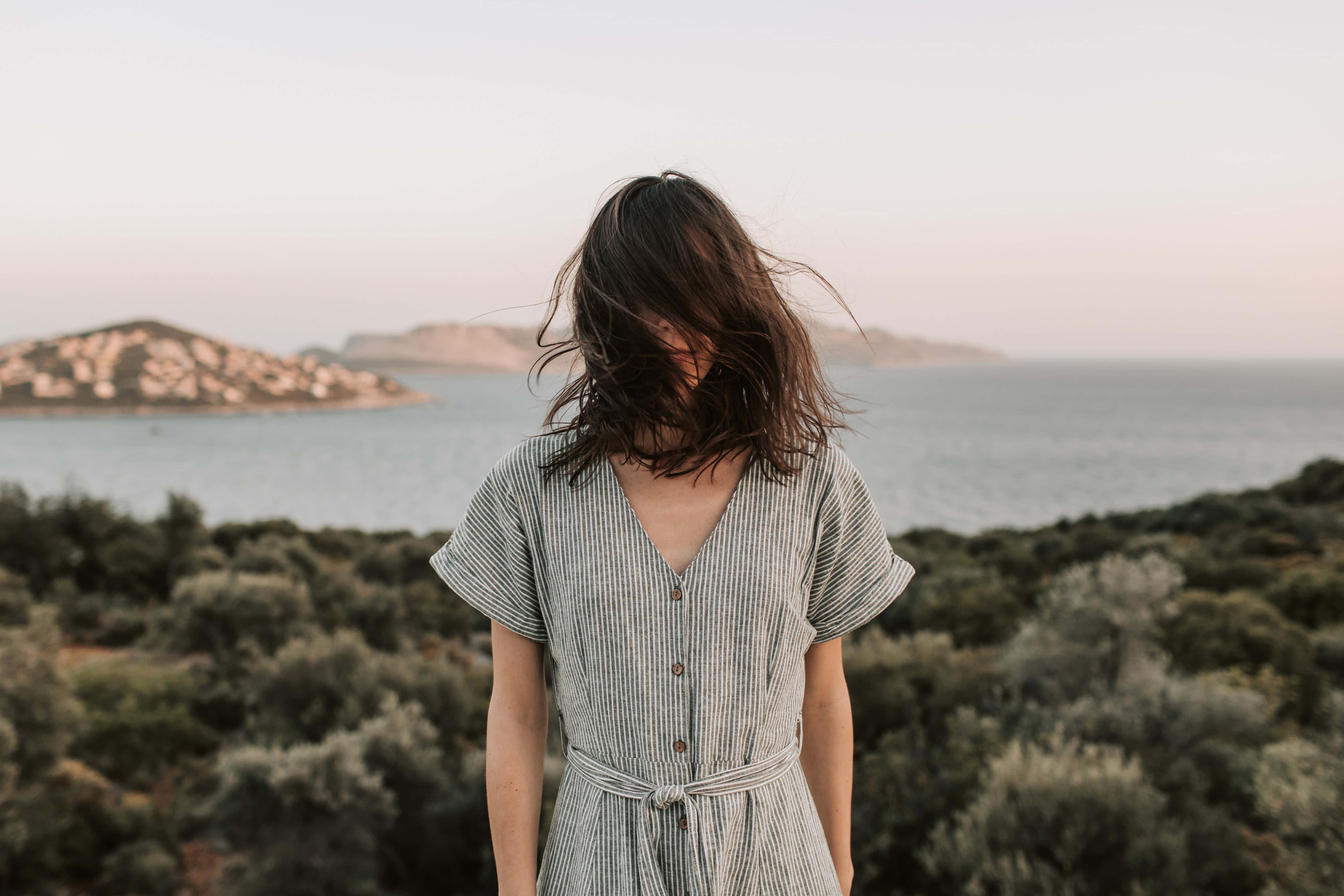 Summer Fashion 桌布 壁紙 — 一位頭髮被風吹動的女性背對鏡頭站立，在日落時分俯瞰平靜的大海和遠處的島嶼。