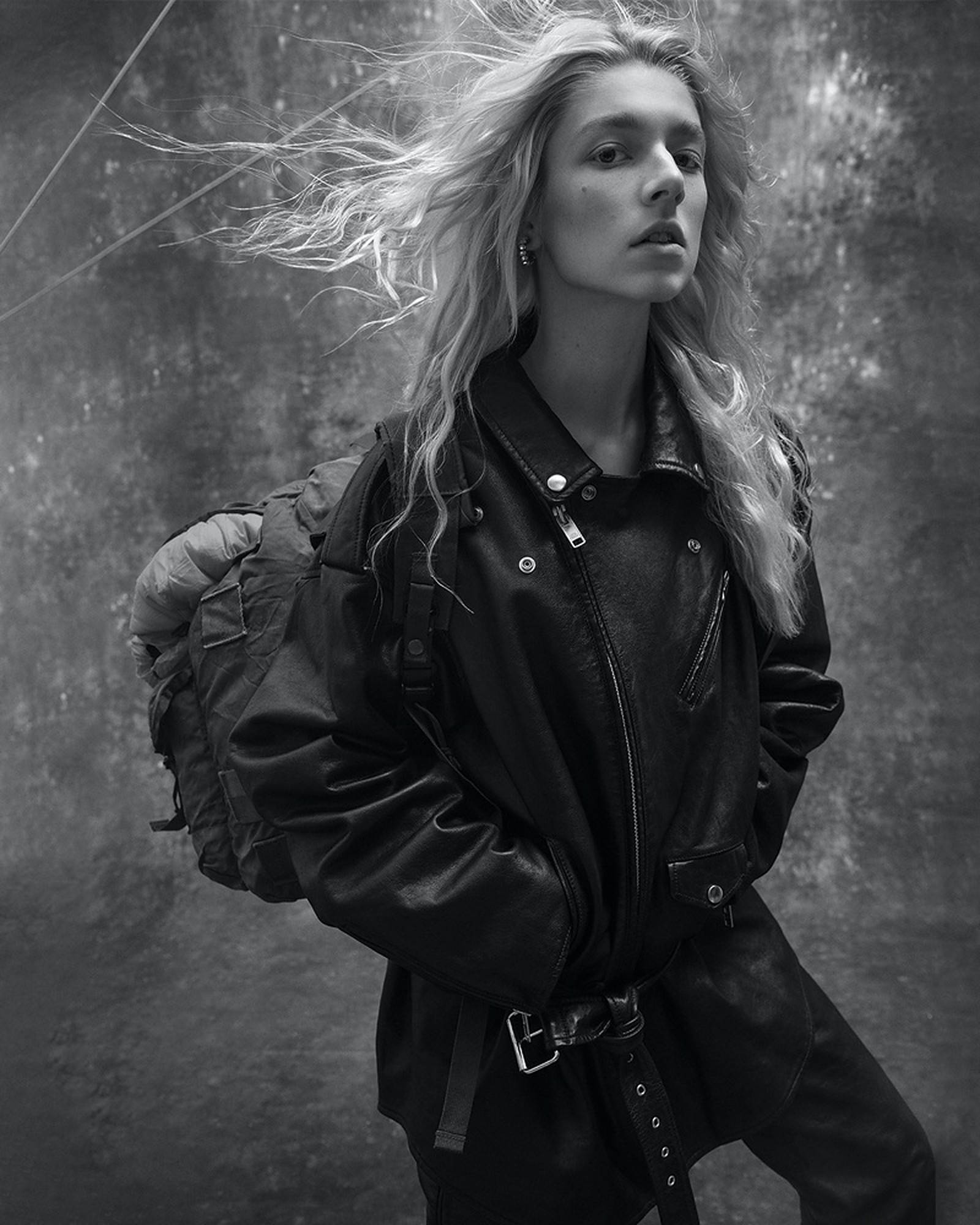 Hunter Schafer 桌布 壁紙 — 一位頭髮被風吹拂的女性，穿著皮夾克和背包，在紋理灰色背景前以單色呈現。