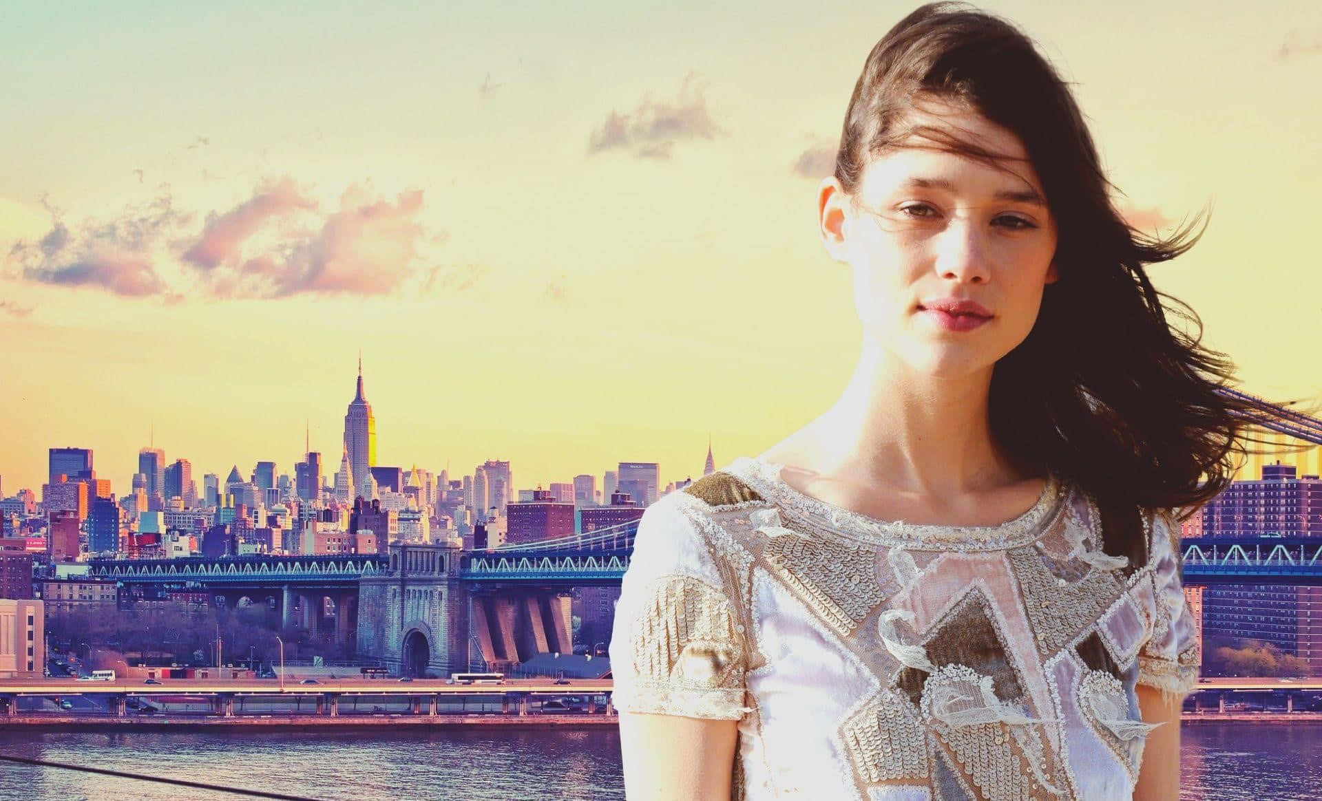 Astrid Berges Frisbey 桌布 壁紙 — 一位頭髮被風吹拂的女性，站在日落時分金黃色的紐約市天際線前。