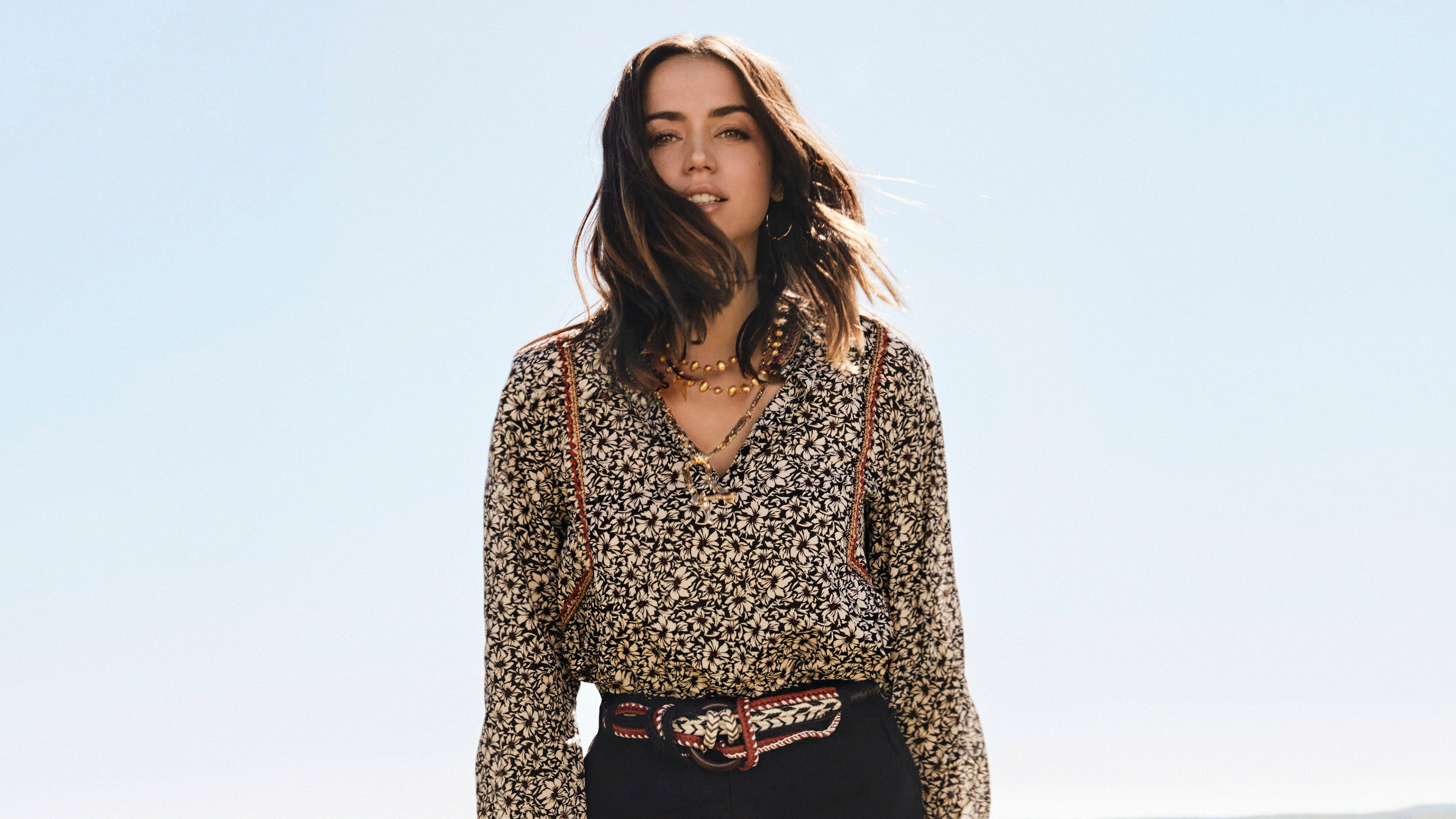 ana de armas 桌布 壁紙 — 一位頭髮被風吹拂的女性，自信地站在清澈的藍天下，穿著圖案襯衫和華麗腰帶。