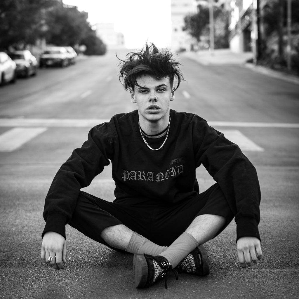 Yungblud 桌布 壁紙 — 一位頭髮豎起的年輕男子在城市街道上盤腿而坐，黑白照片中直視鏡頭。