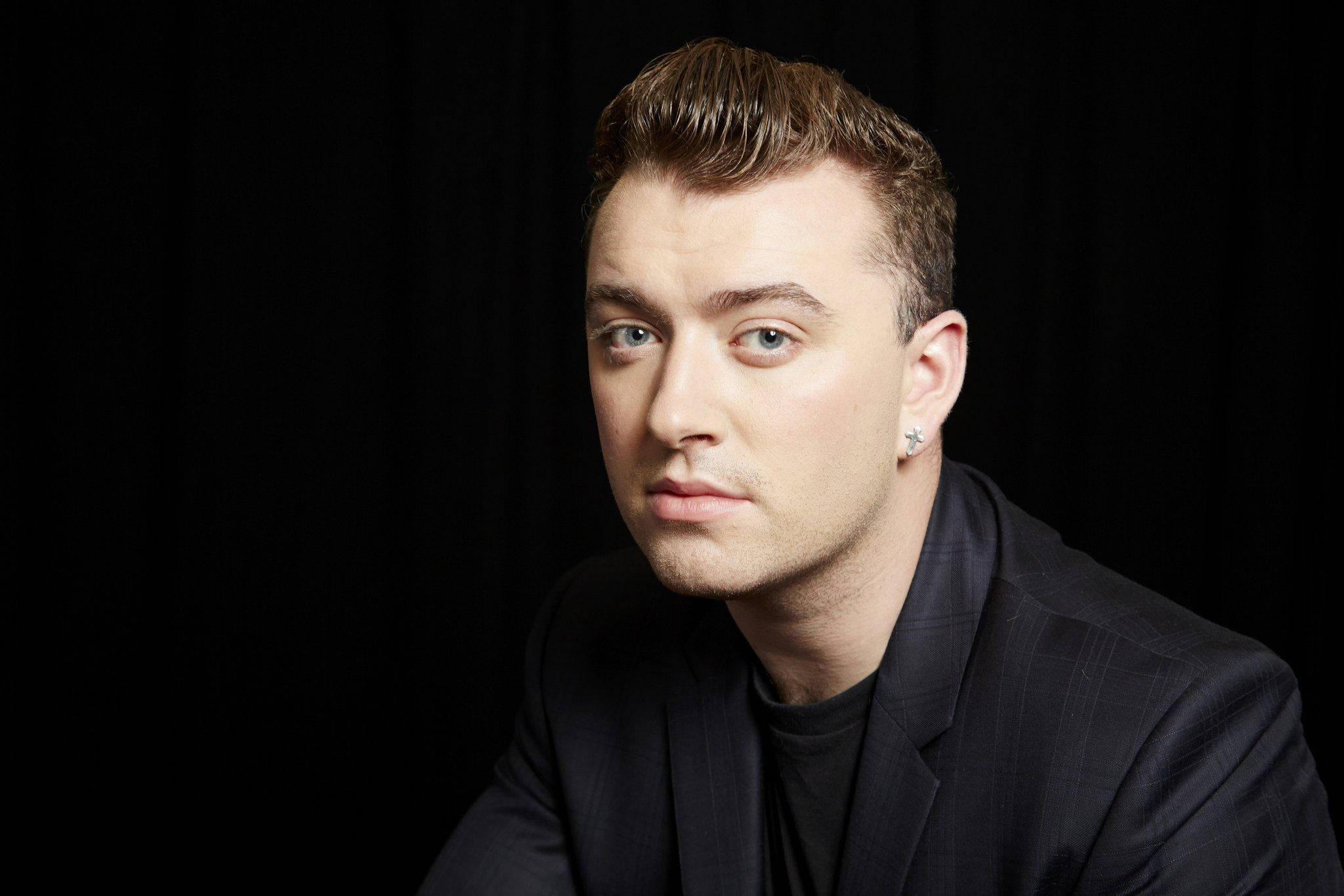 Sam Smith 桌布 壁紙 — 一位頭髮造型時尚、戴著星星耳環的男子，身穿俐落西裝，在純黑背景下凝視前方。