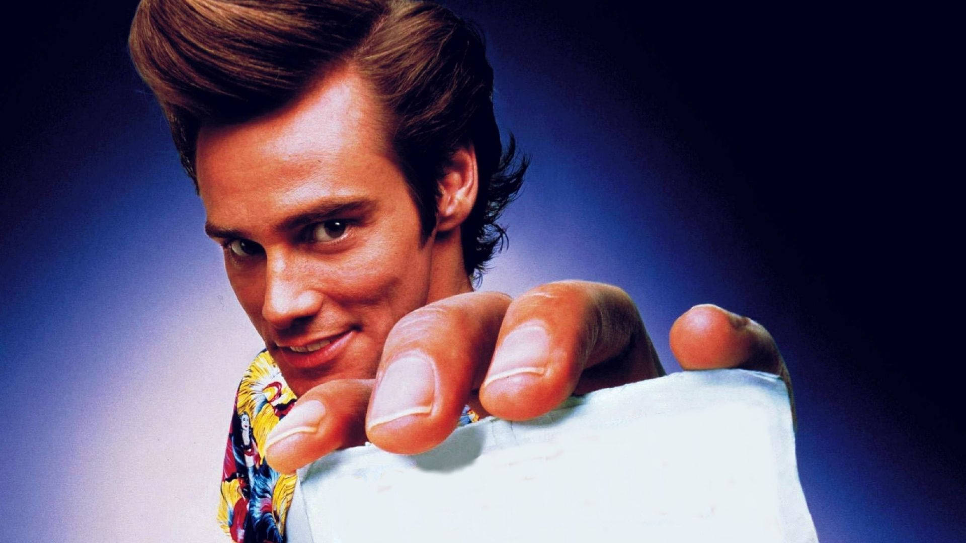 Jim Carrey 桌布 壁紙 — 一位頭髮造型時尚、身穿彩色襯衫的男子，在藍色漸層背景前向觀眾展示一件白色物體。