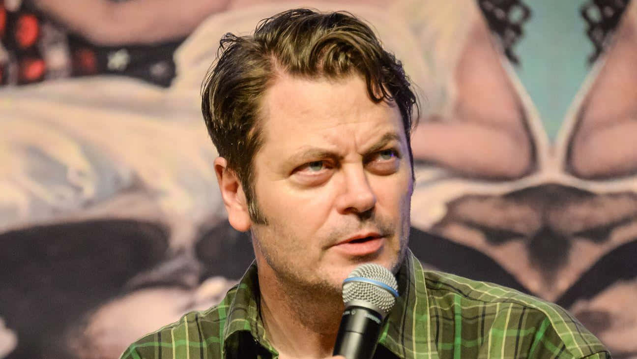 nick offerman 桌布 壁紙 — 一位頭髮造型時尚的男子，穿著綠色格紋襯衫，在模糊的壁畫背景下對著麥克風說話。
