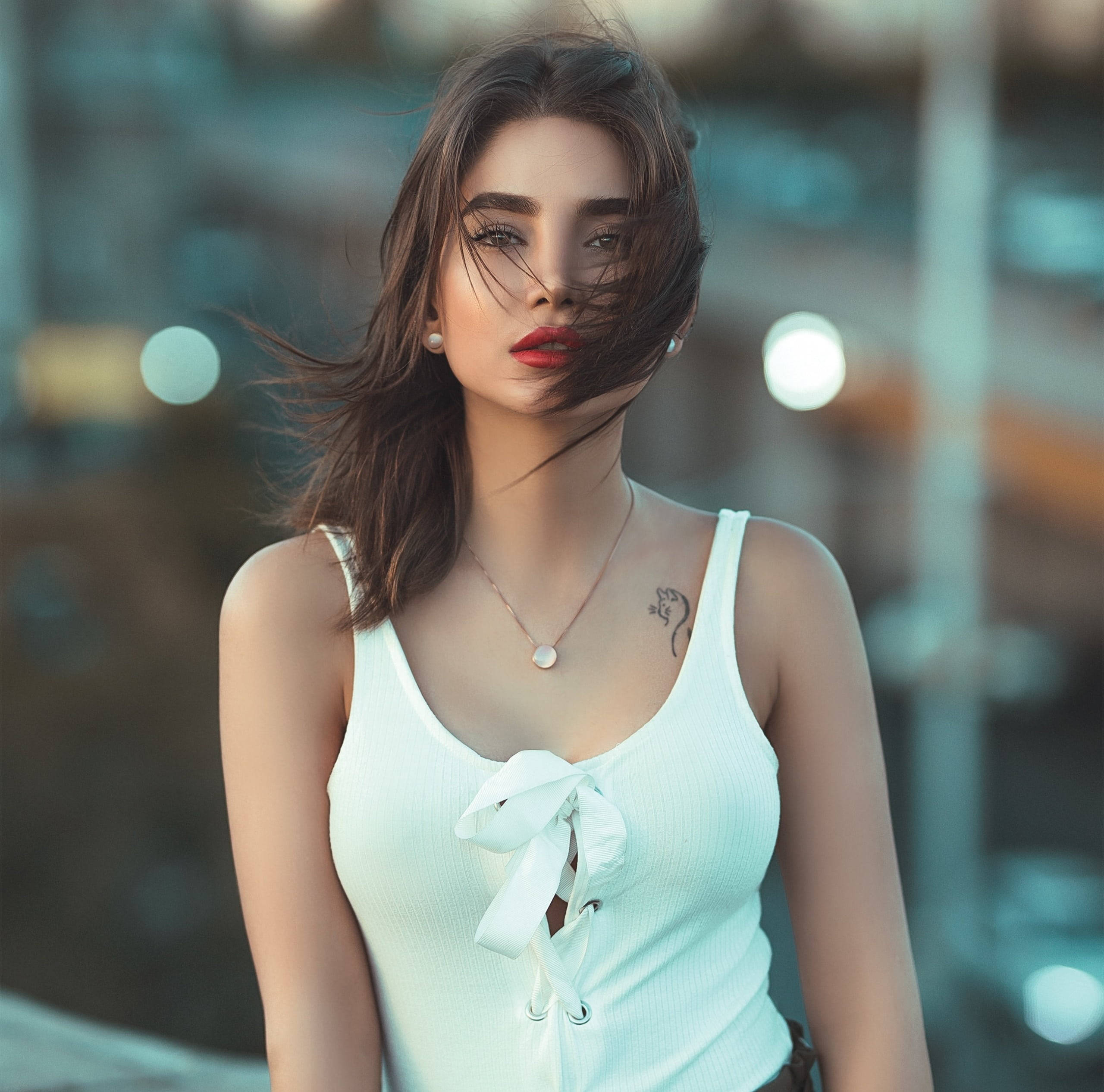 美女 桌布 壁紙 — 一位頭髮隨風飄揚、嘴唇鮮紅大膽的女性，自信地穿著白色羅紋上衣，背景是模糊的城市景觀。