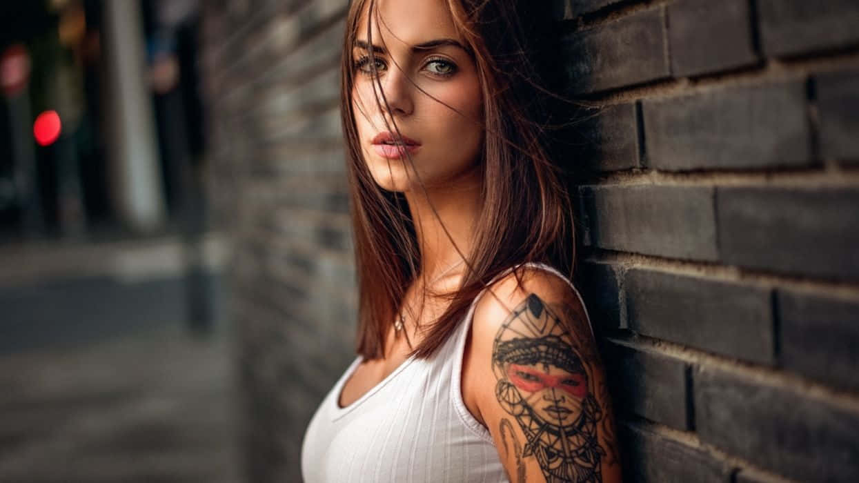 Tattoo 桌布 壁紙 — 一位頭髮隨風飄揚、帶有部落紋身的女性，在城市小巷中靠著深色磚牆。