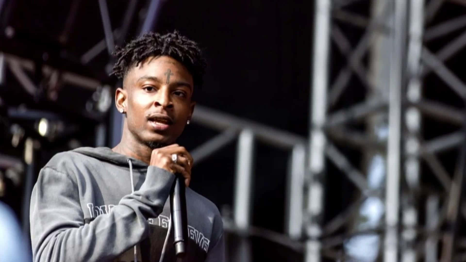 21 Savage 桌布 壁紙 — 一位額頭上有十字刺青的年輕男饒舌歌手在舞台燈光下熱情地對著麥克風表演。