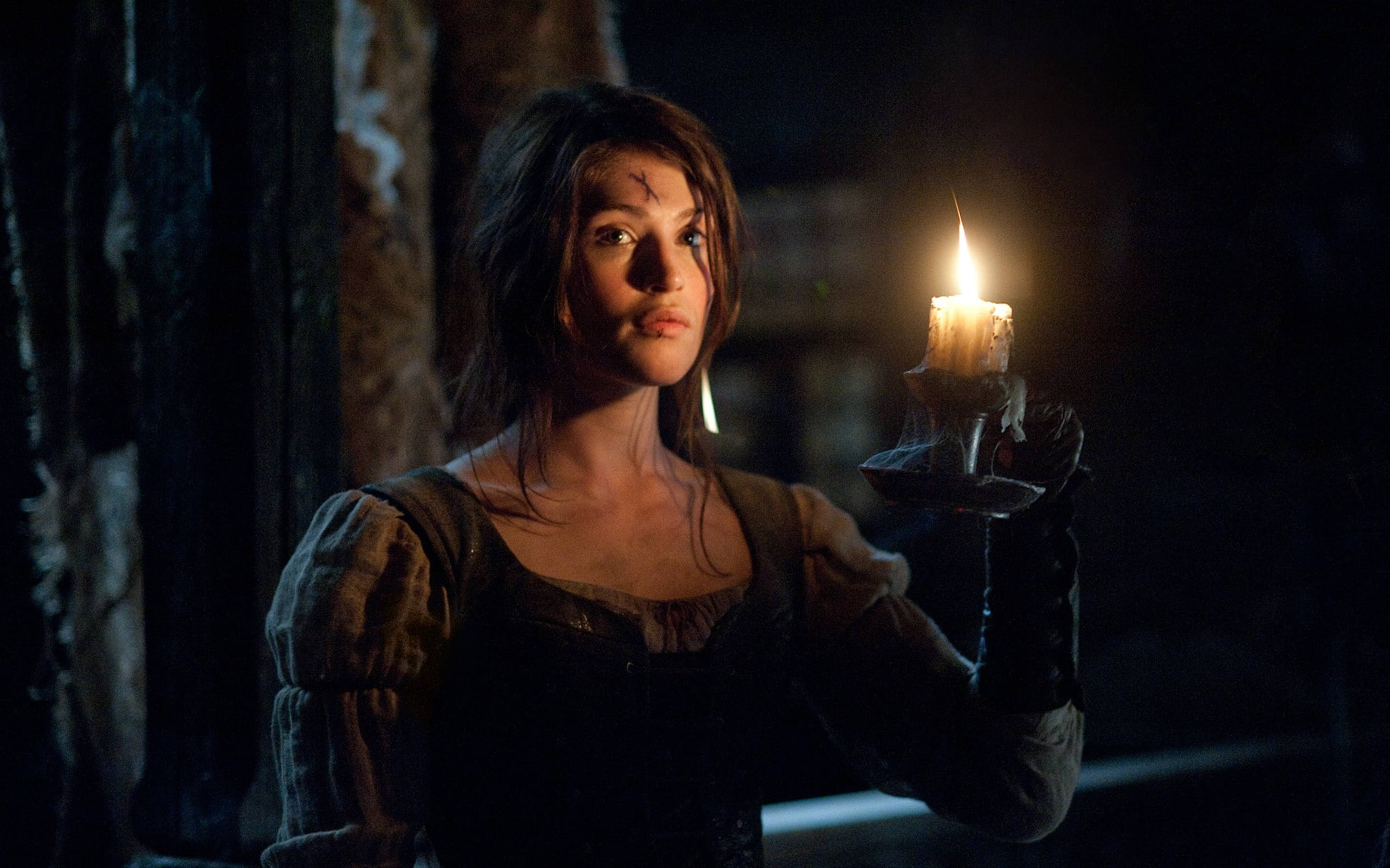 Gemma Arterton 桌布 壁紙 — 一位額頭有疤痕的女性在黑暗的鄉村風格房間中手持一支閃爍的蠟燭，溫暖的光芒映照在她的臉上。
